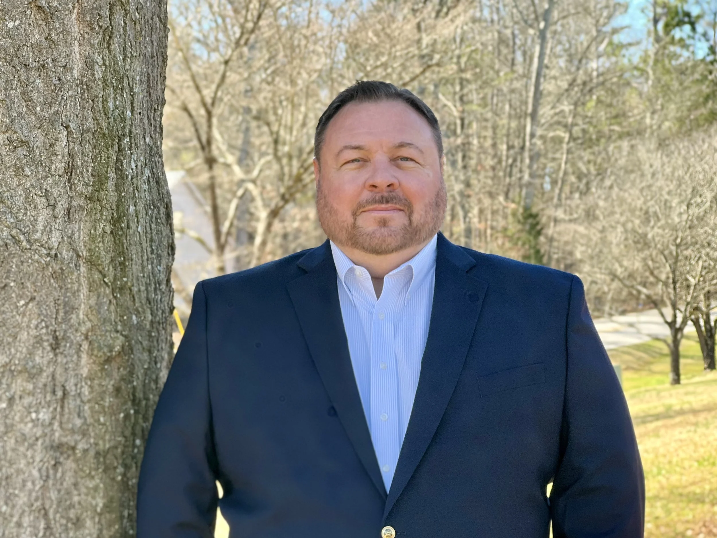 A smiling man with glasses, facial hair, and a checked blue shirt standing outdoors next to a tree with blurred trees and sky in the background.