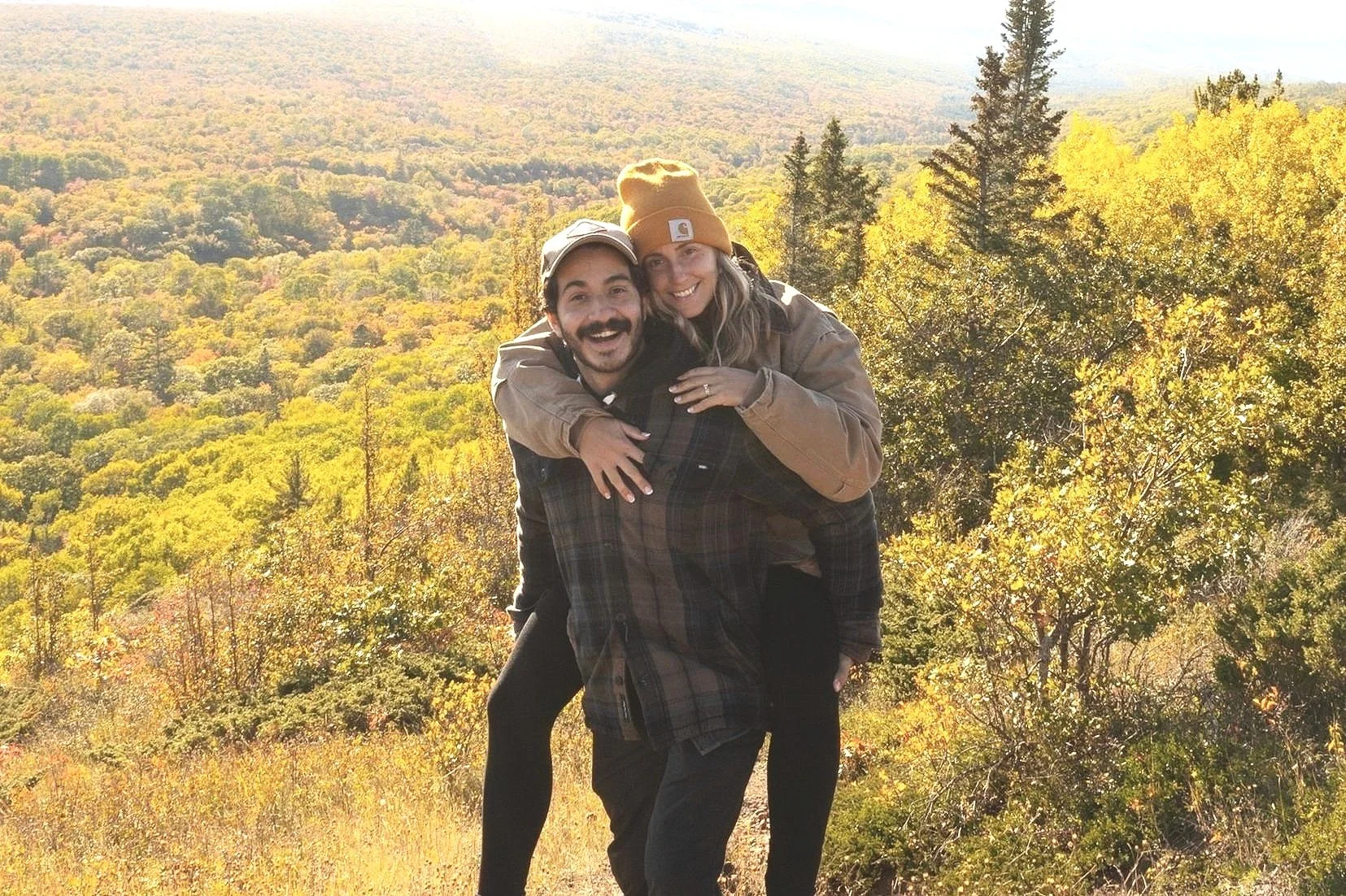 A man and woman smiling and hugging outdoors in a forested area with autumn foliage.