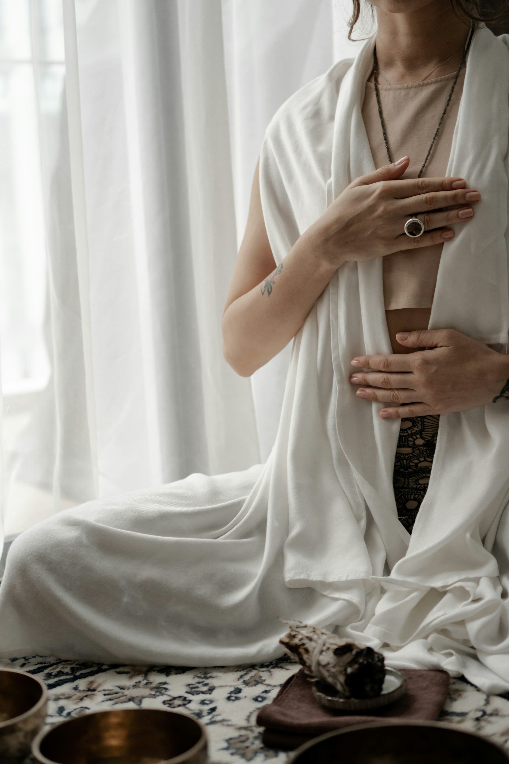 A woman sitting on a patterned rug near a window, wearing a white silk or satin robe with a beige top underneath. She has tattoos on her left arm and right wrist, and is holding her chest and abdomen with her hands. She is wearing a ring and a necklace. There are bowls and a textured object on a small cloth in front of her.