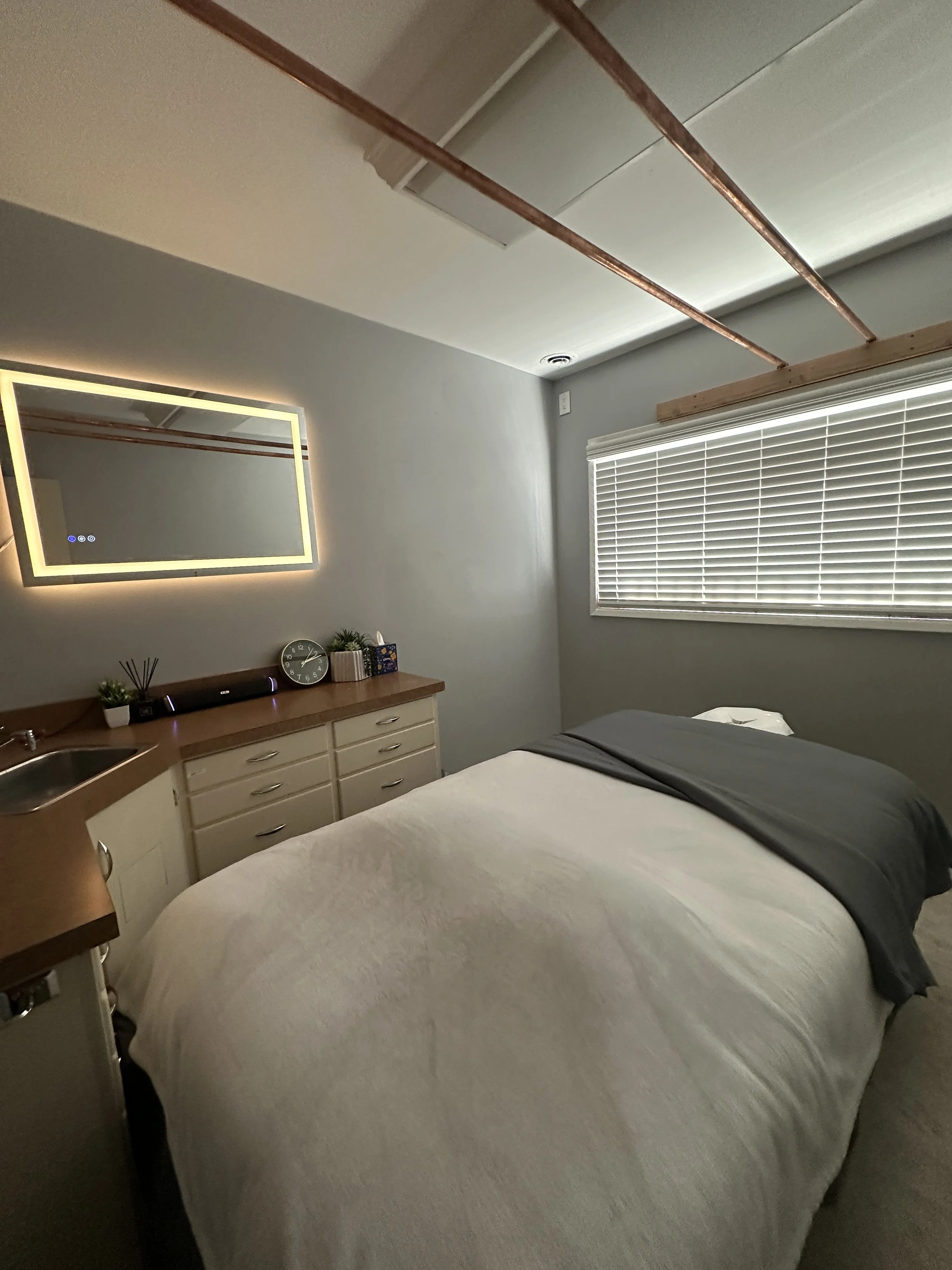 A bedroom with a bed covered in white and gray bedding, a window with blinds, a dresser with a clock, potted plants, and decorations, a mirror with LED lighting, and exposed wooden ceiling beams.