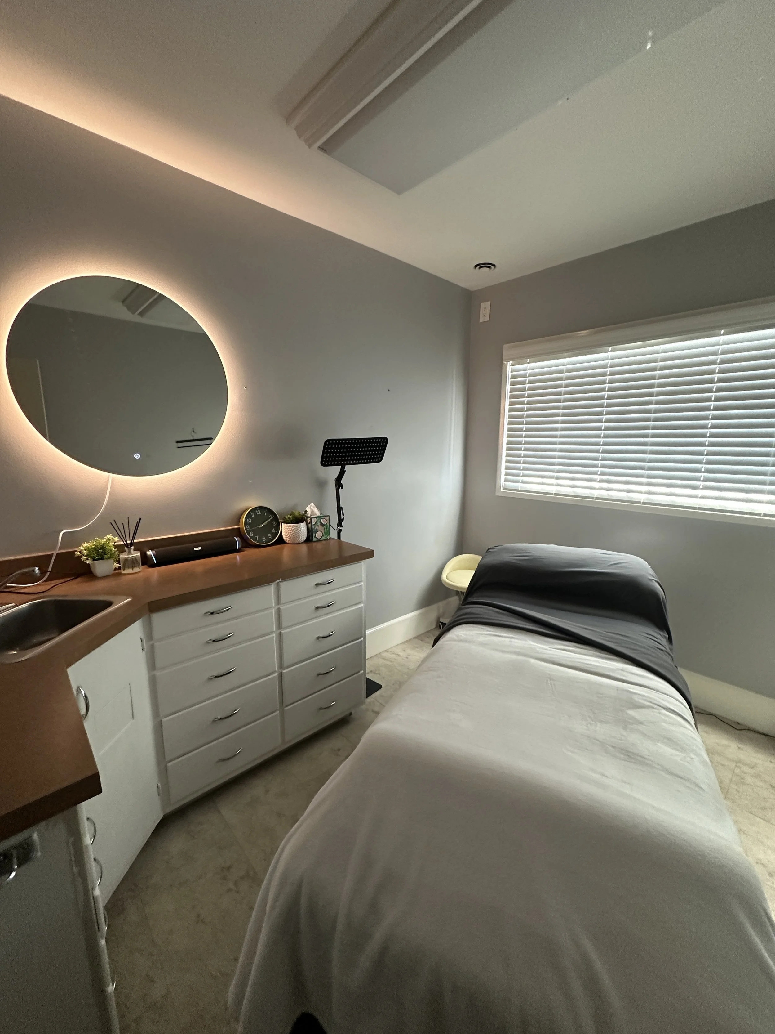 A massage room with a massage table draped in a white sheet, positioned near a window with blinds. There is a cabinet with plants, a clock, and a music speaker, and a circular mirror with backlighting on the wall.
