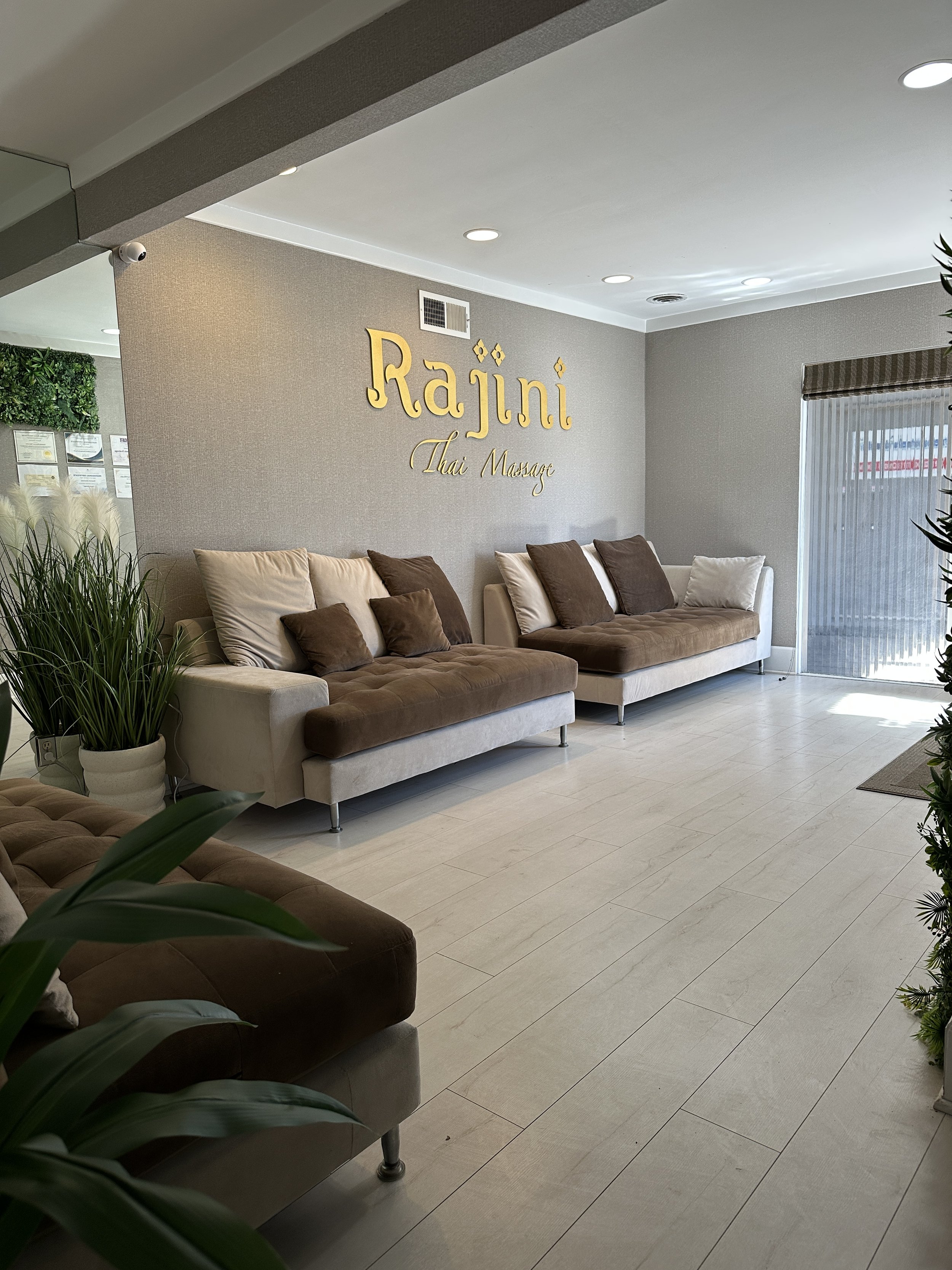 Waiting area in a Thai massage spa with beige and brown couches, green plants, and a sign on the wall that says 'Rajini Thai Massage'.
