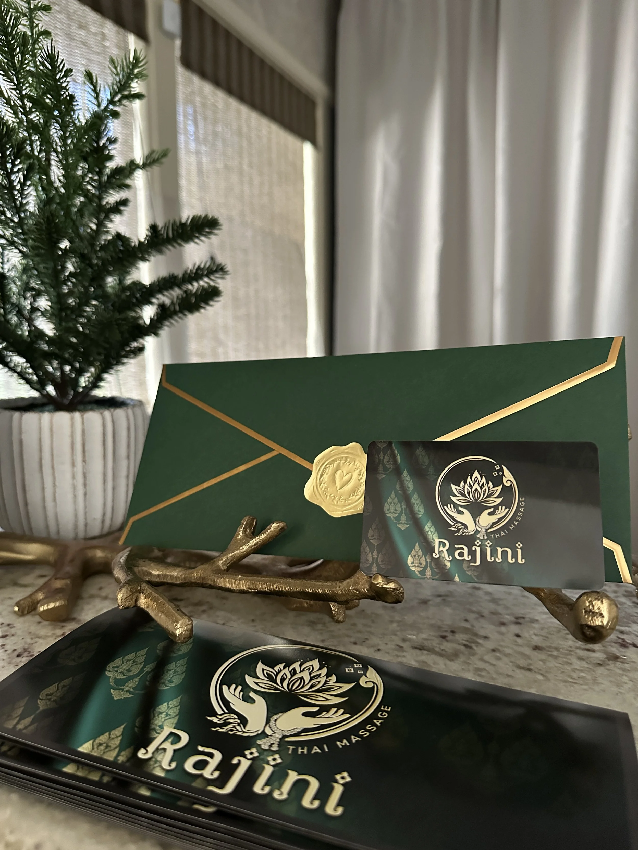 An advertisement for Rajini Thai Massage gift certificates, depicting a logo with a lotus flower, green and gold text, and yellow stars, promoting gift cards as perfect presents for any occasion.