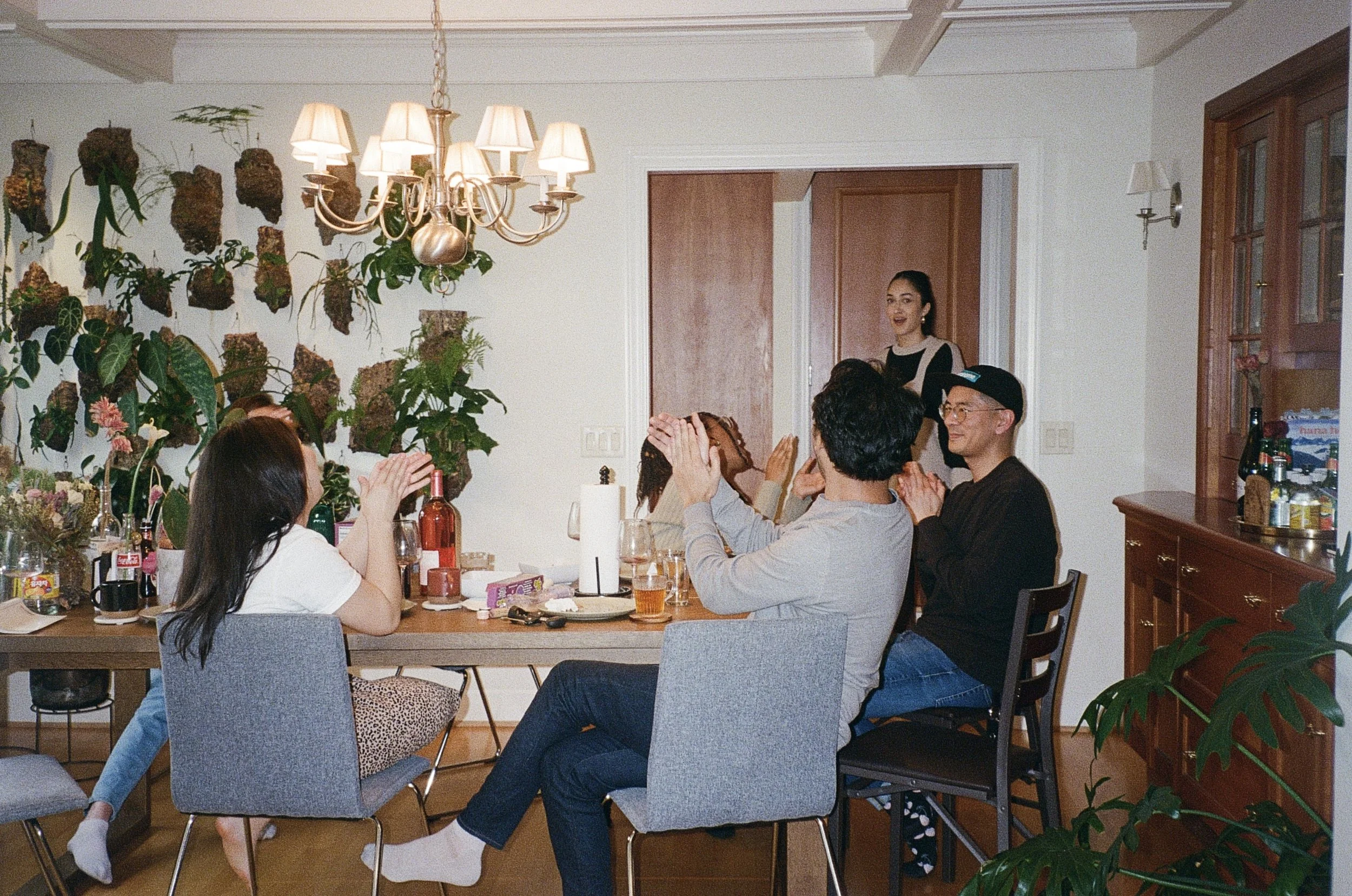 Group of people celebrating at a dinner table with a girl covering her face, others clapping, in a warmly lit dining room with plants and wooden furniture.