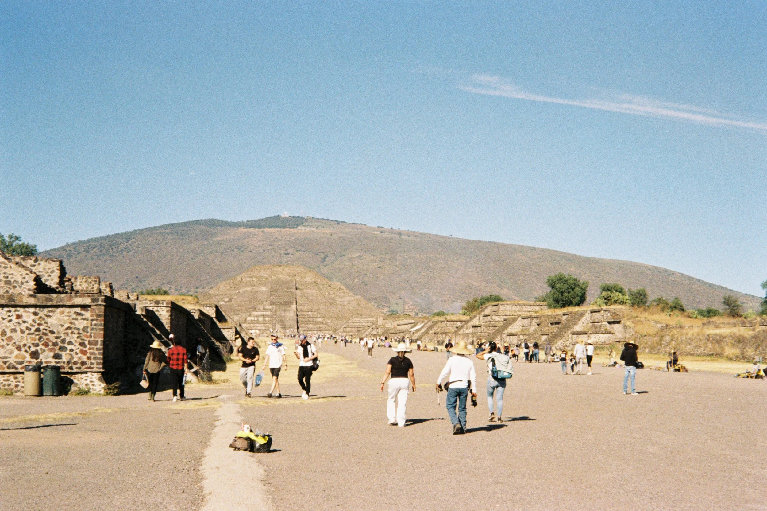 Tourists walking around the ancient ruins of Teotihuacan with pyramids and stone structures under a clear blue sky.