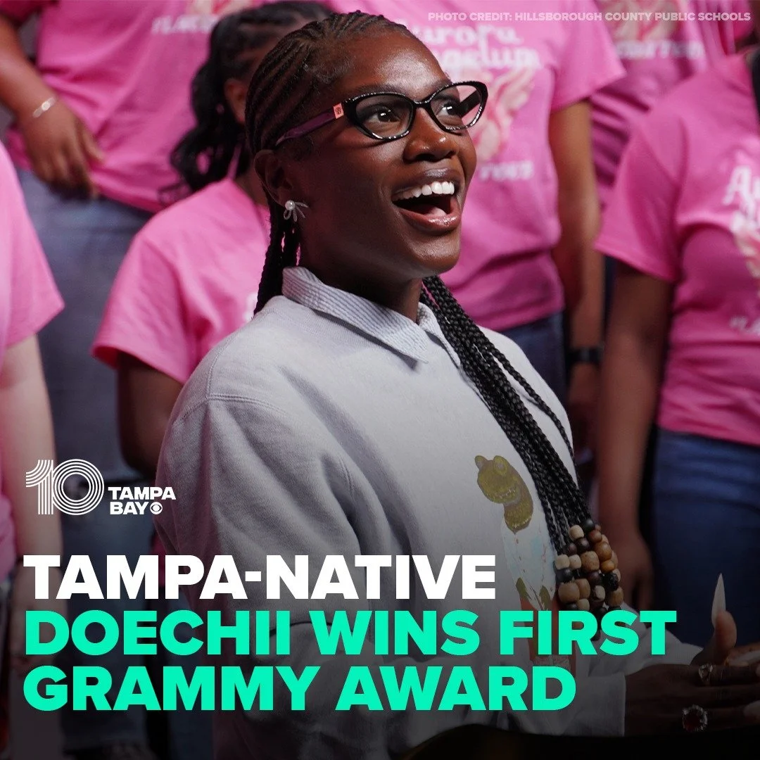 A woman with glasses and braided hair, smiling and appearing happy among a group of people wearing pink shirts, celebrating her winning a Grammy award.
