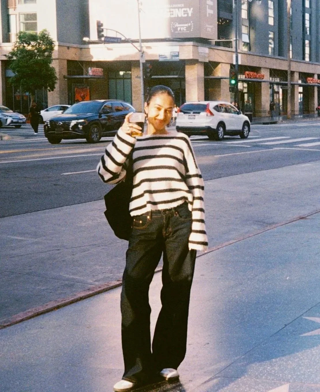 A woman standing on a city sidewalk, smiling and holding a drink, wearing a black and white striped sweater and black pants.