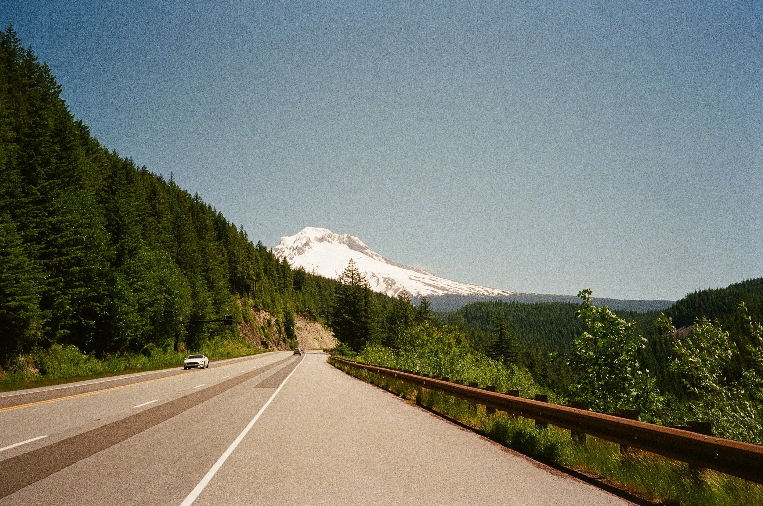 A scenic road winding through lush green forested hills with Mount Hood snow-capped mountain in the background, clear blue sky overhead, and a few cars driving along the road.