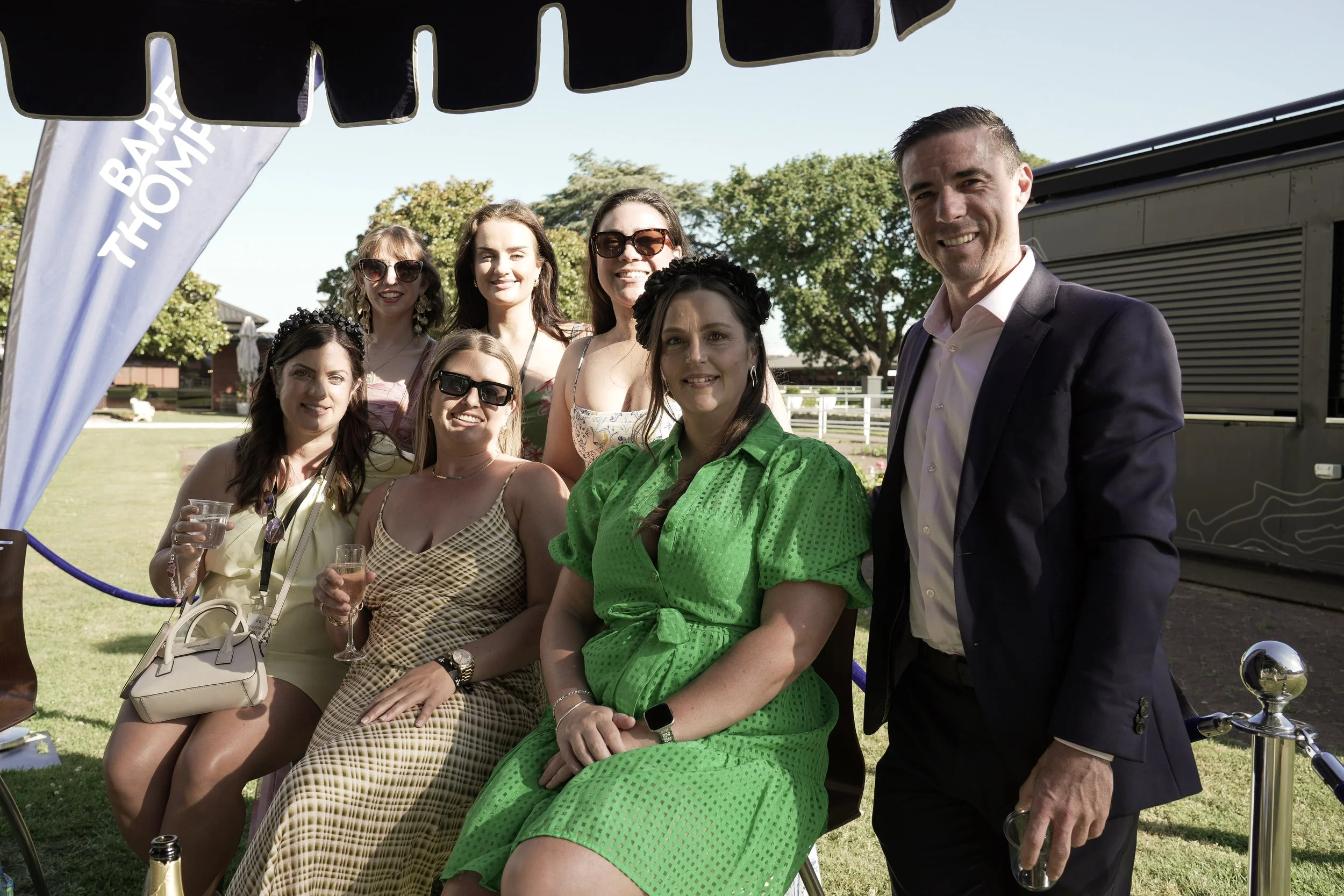 Group of nine people, eight women and one man, gathered outdoors at a sunny event. Some women are sitting, and the man is standing. Several hold glasses of drinks, and everyone is smiling. The women are dressed in colorful summer dresses, and the man