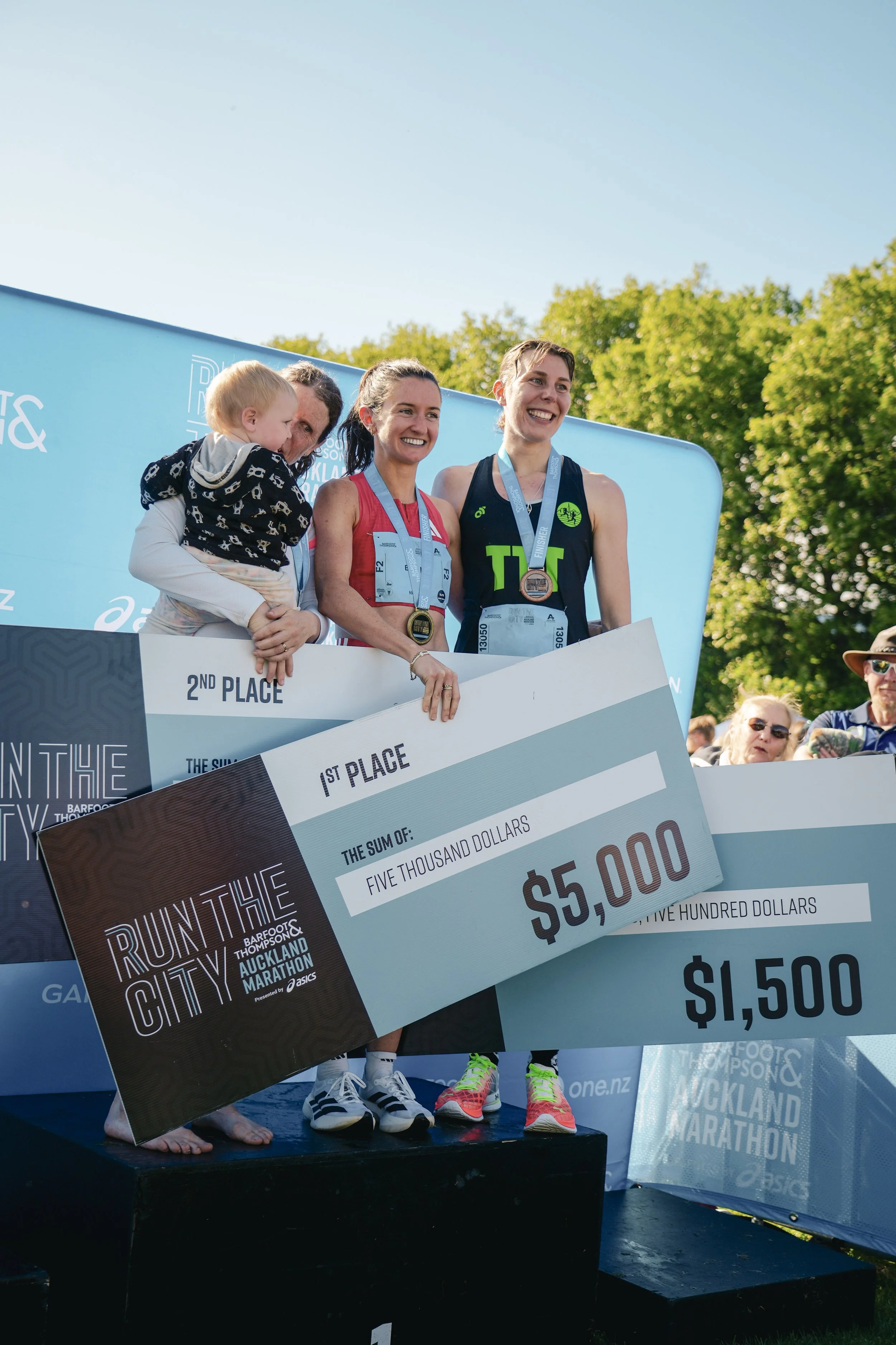 Three women standing on a podium holding oversized checks and medals, celebrating at a marathon event, with spectators in the background and greenery under a clear sky.