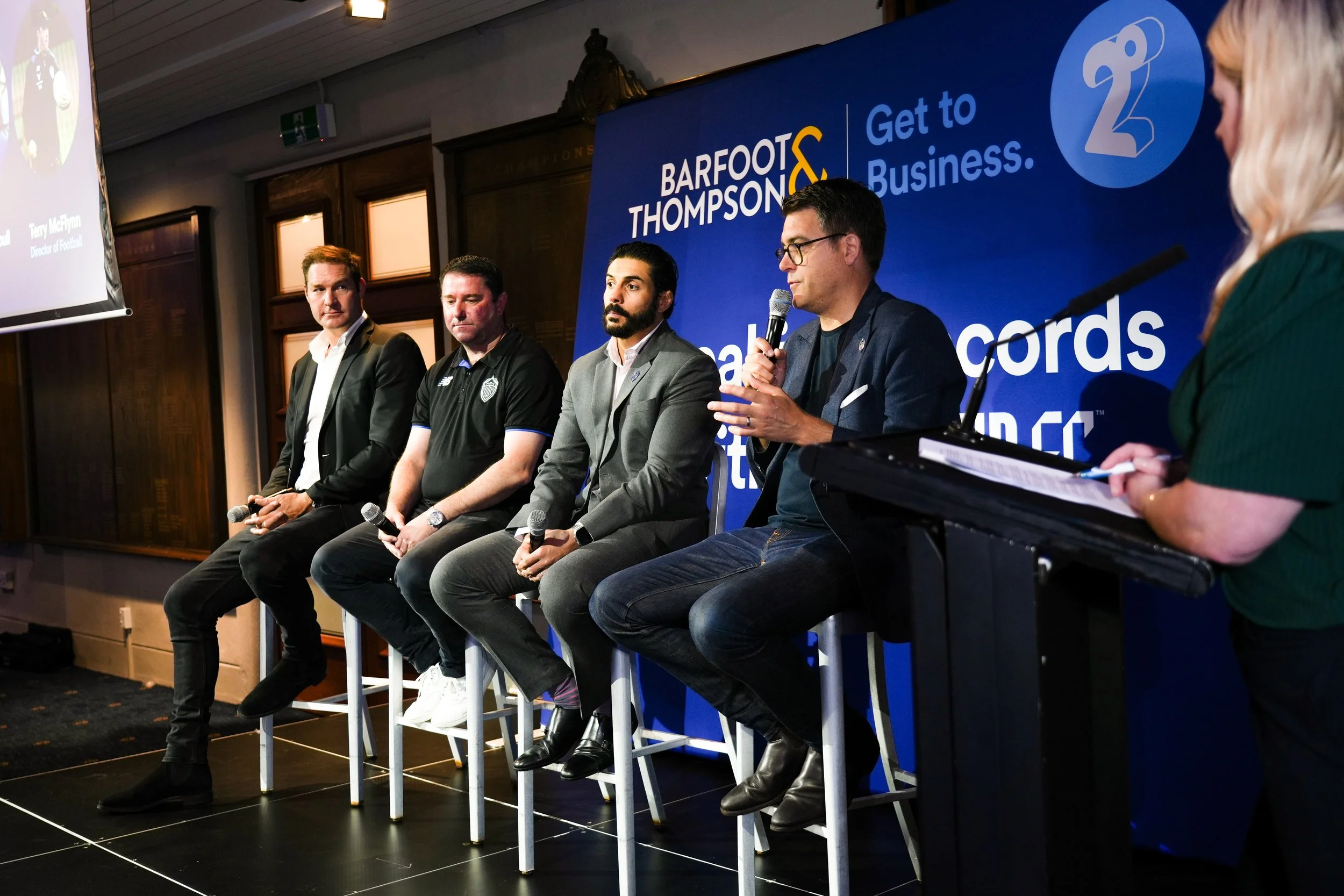 Four men sitting on stools on a stage during a panel discussion, with a woman standing at a podium on the right. Behind them is a blue backdrop with the text 'Barfoot & Thompson' and a logo. One man is speaking into a microphone, and the woman appear