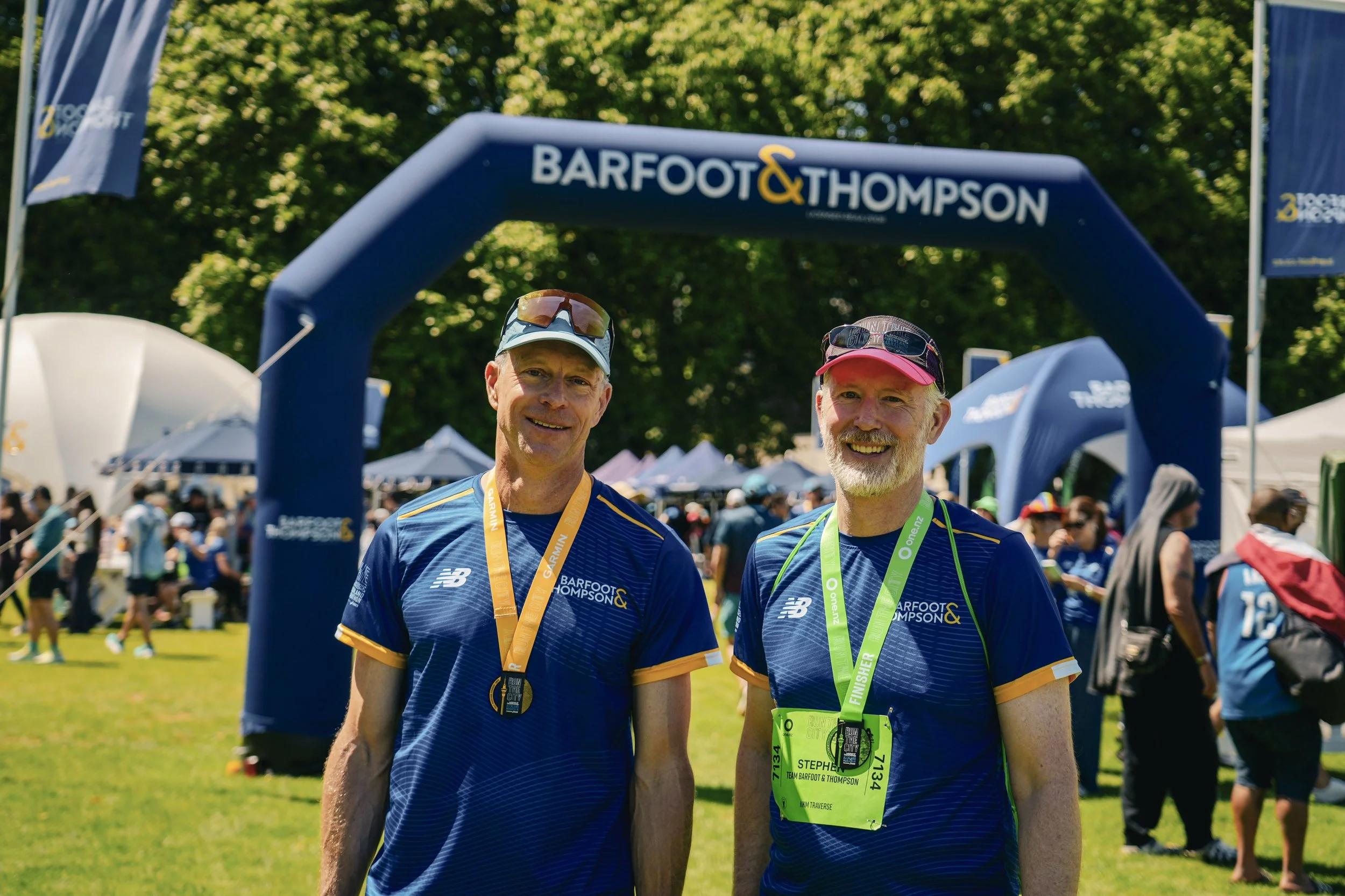 Two men wearing blue marathon shirts and medals around their necks smiling in front of a blue arch that says 'Barfoot & Thompson' at a race event.