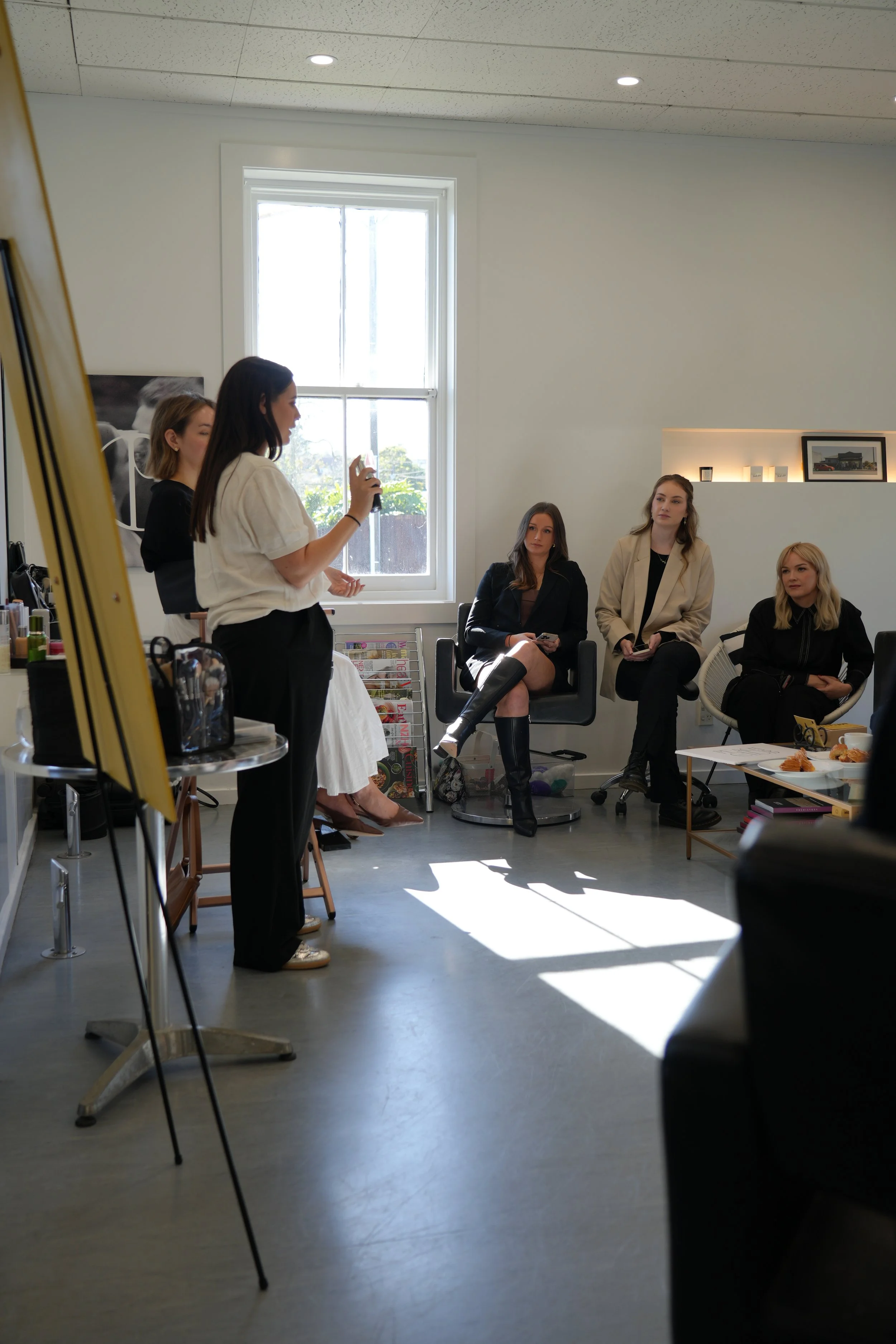Four women in a meeting or workshop, with one standing and speaking, three seated listening, in a bright room with a large window, and various items on tables.