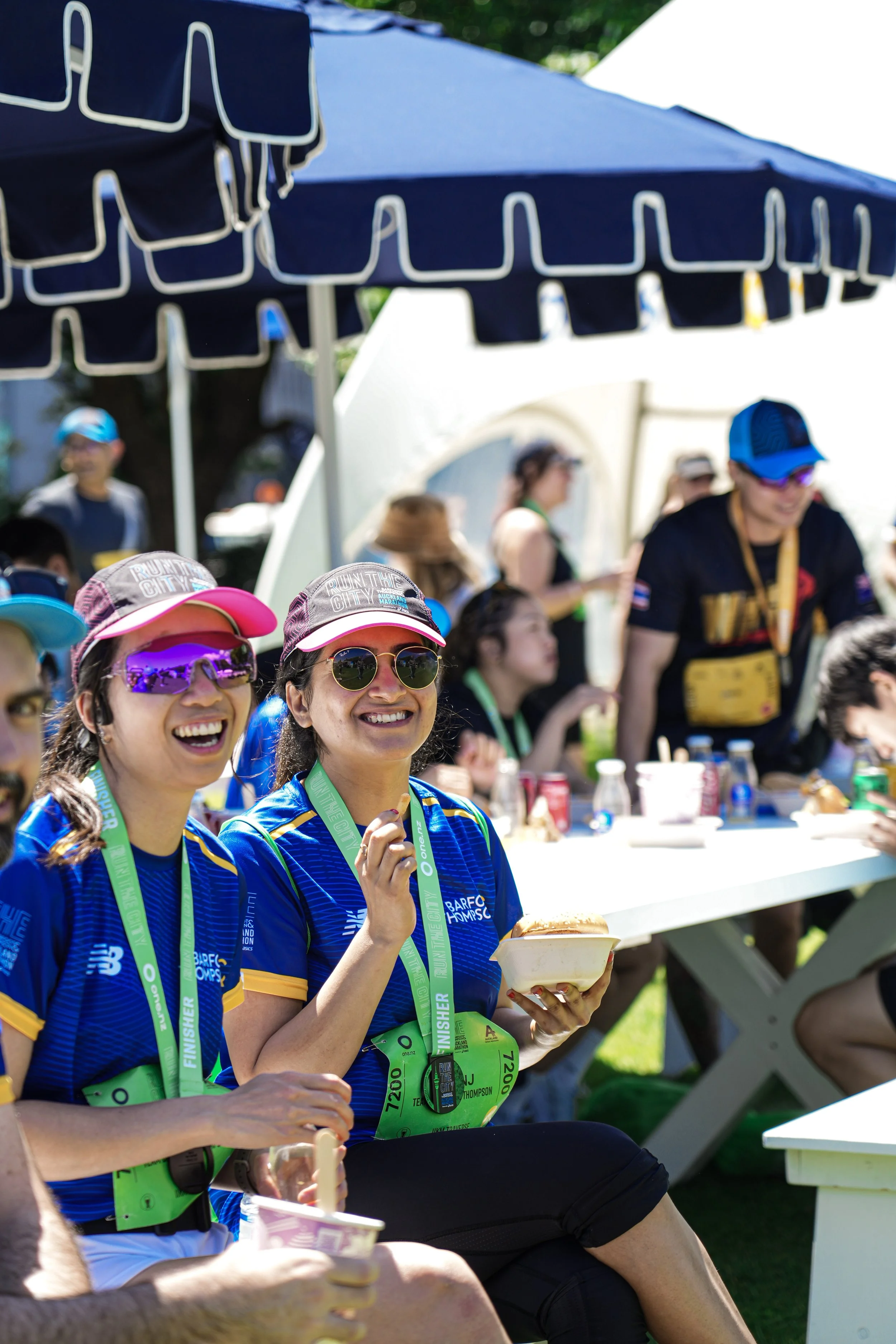Two women in blue running shirts and medals around their necks smiling, sitting near a table with food and drinks, at an outdoor event under a blue canopy.