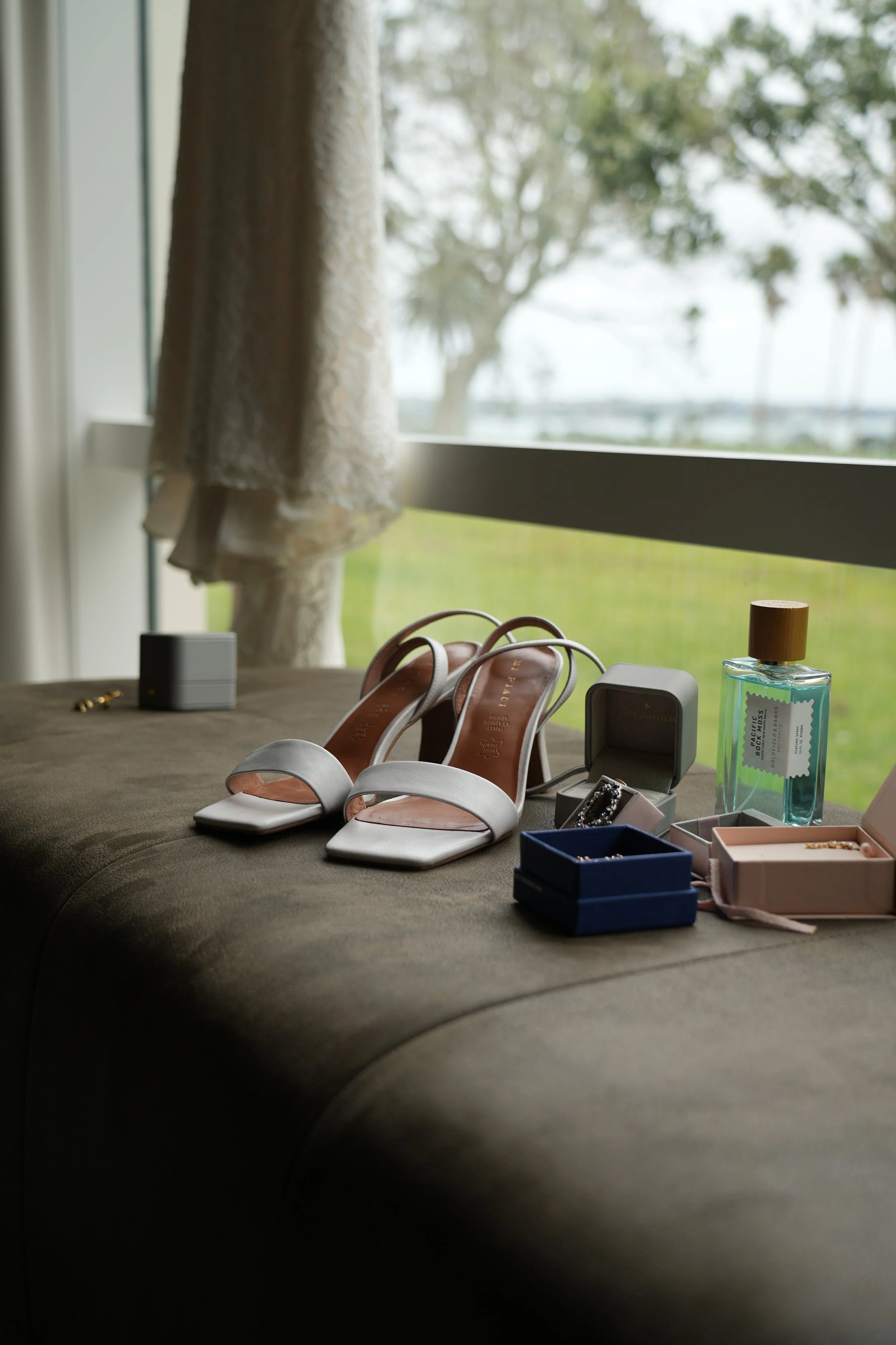 A pair of white open-toe heels, jewelry boxes, perfume, and jewelry placed on a cushioned surface near a window with a view of green grass and trees outside.