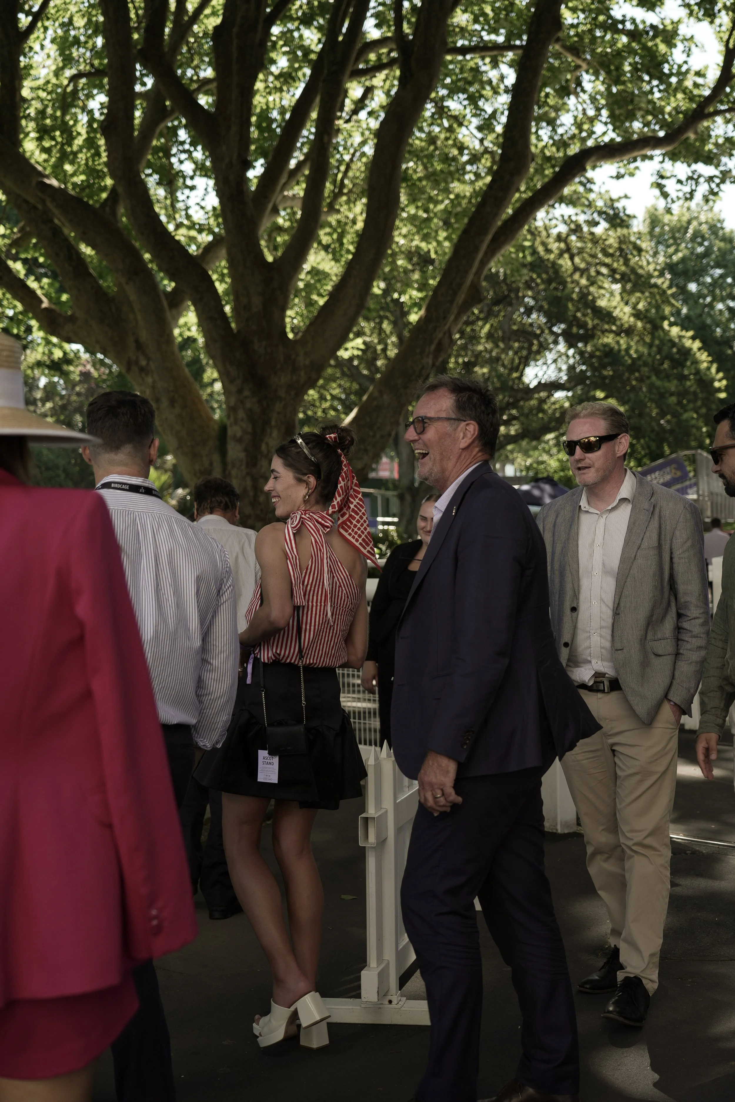 A group of people socializing outdoors under a large leafy tree. The people are dressed in smart casual and summer attire, and some are smiling and laughing.