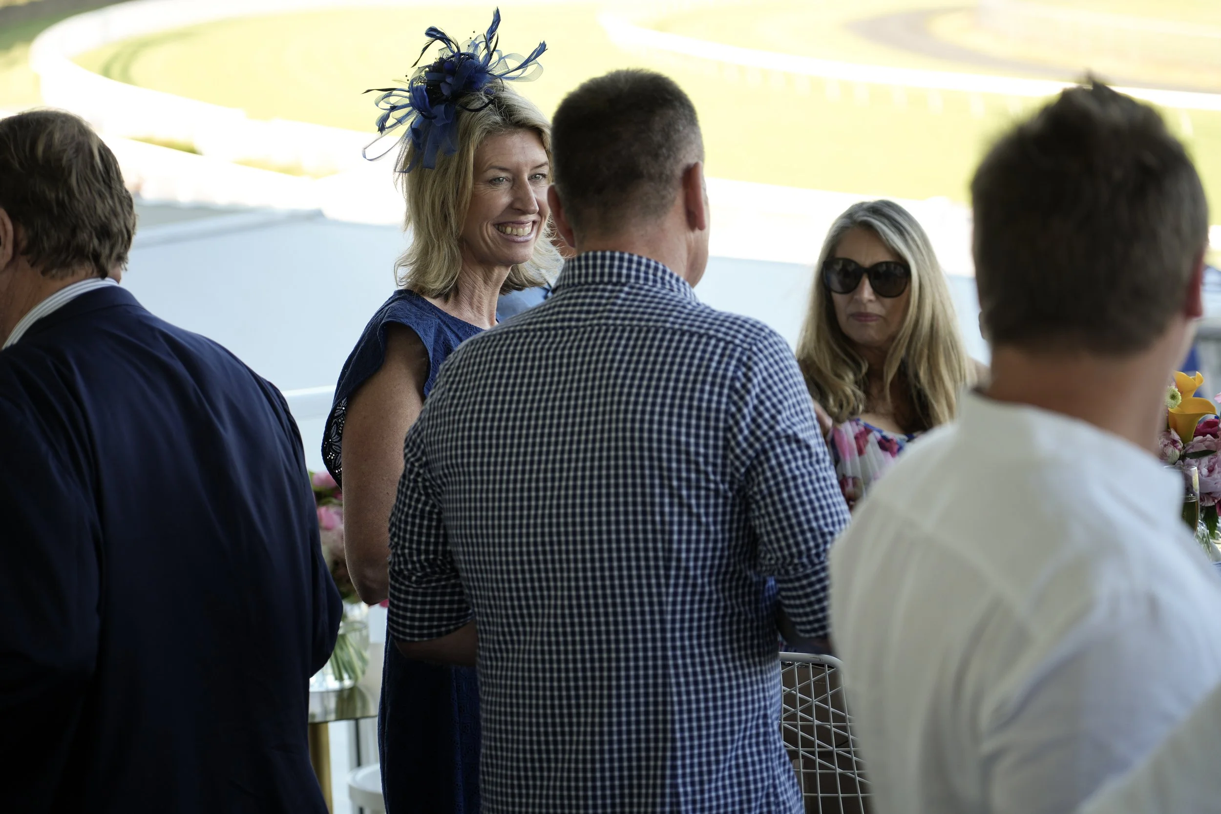 Group of five people standing and conversing outdoors, with a woman wearing a blue dress and a blue fascinator smiling, and a woman wearing sunglasses on the right.