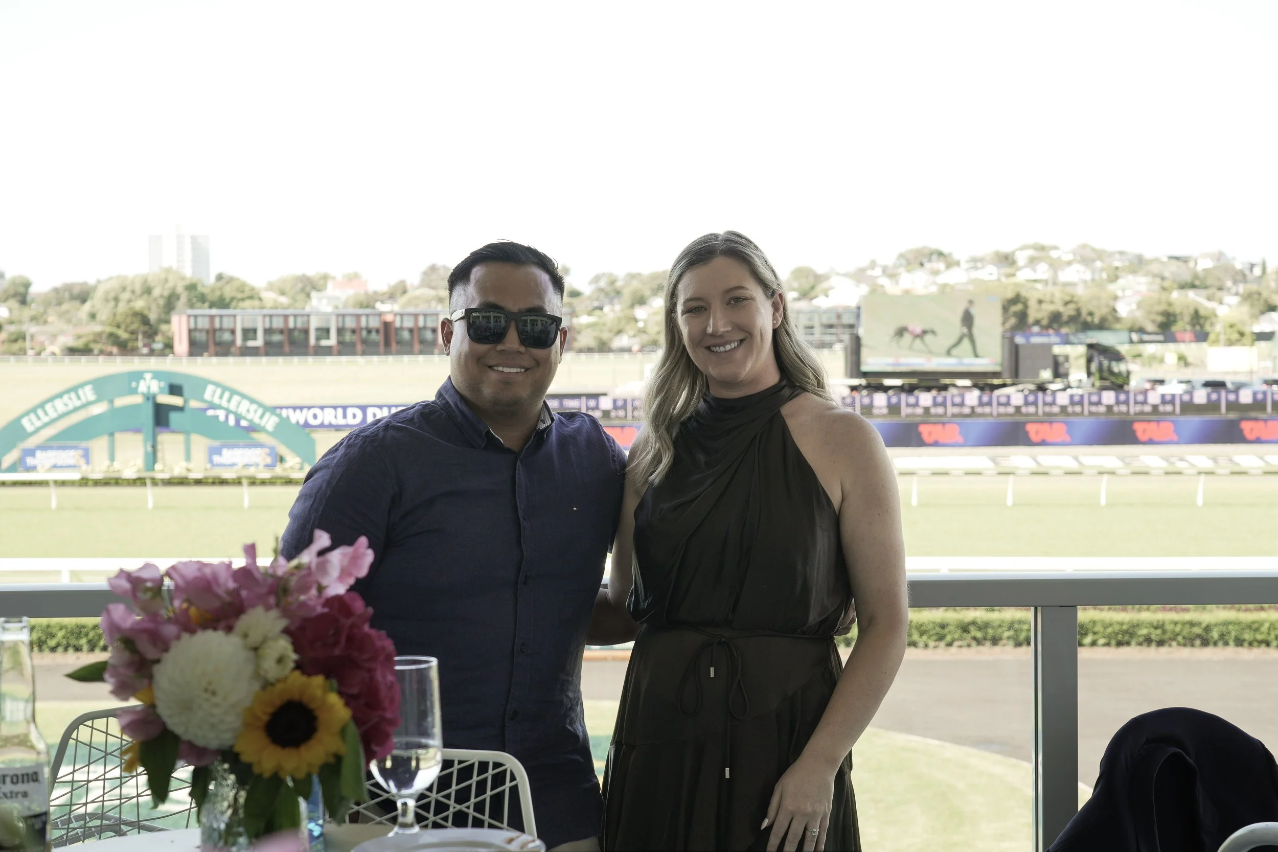 A man and woman smiling at the camera at a terrace overlooking a horse racing track with a large electronic screen in the background.