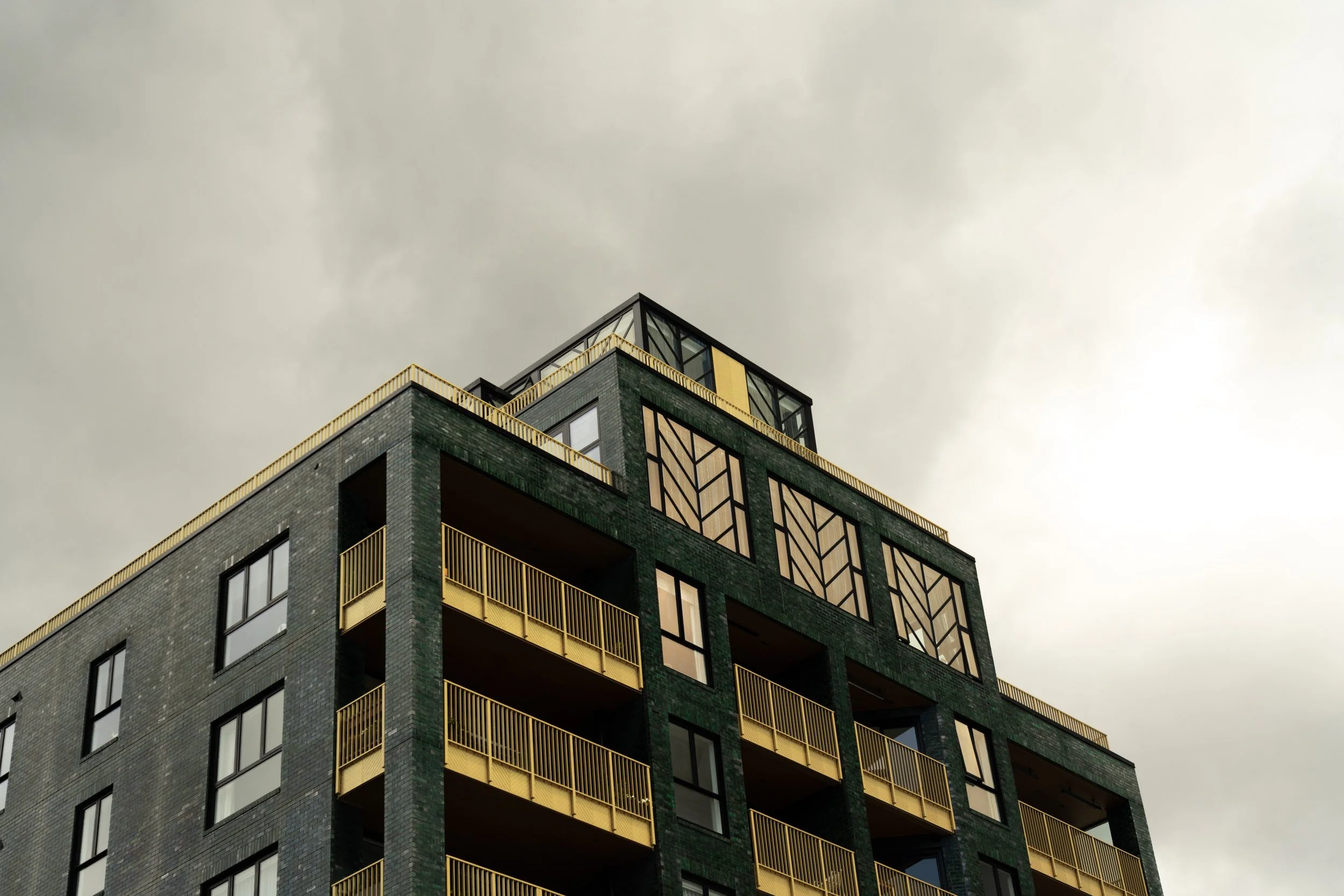 A modern multi-story building with dark gray brick exterior, yellow railings on balconies, and decorative window panels, under a cloudy sky.