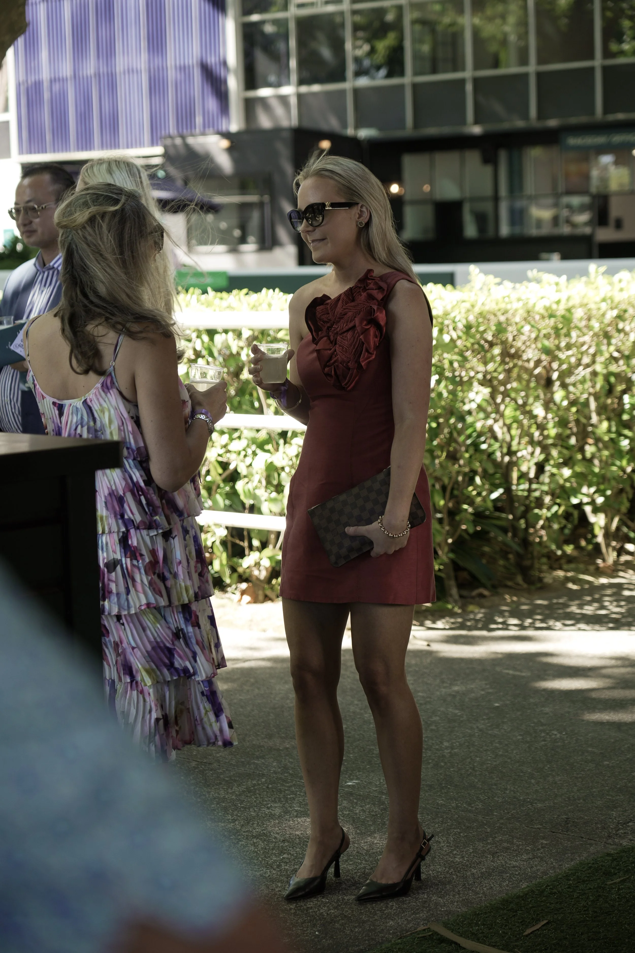 A woman in a red dress with sunglasses and high heels holding a clutch purse, standing outdoors, talking to another woman holding a drink, with other people and greenery in the background.