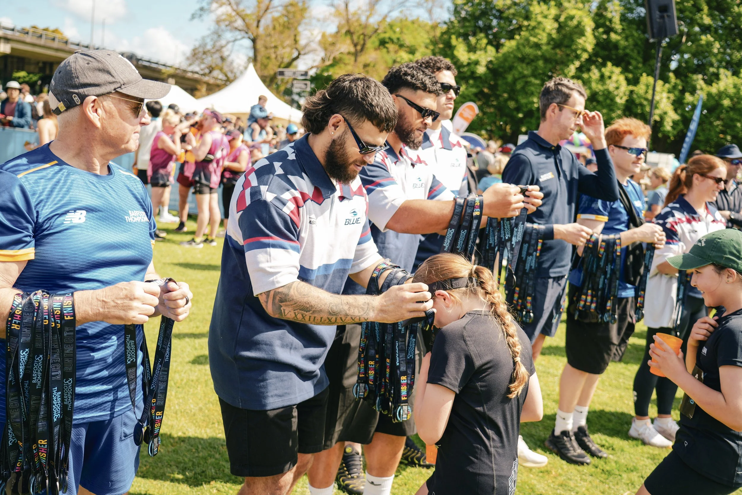 Group of people, some wearing sports jerseys, receiving medals at an outdoor event on a grassy field with trees and tents in the background