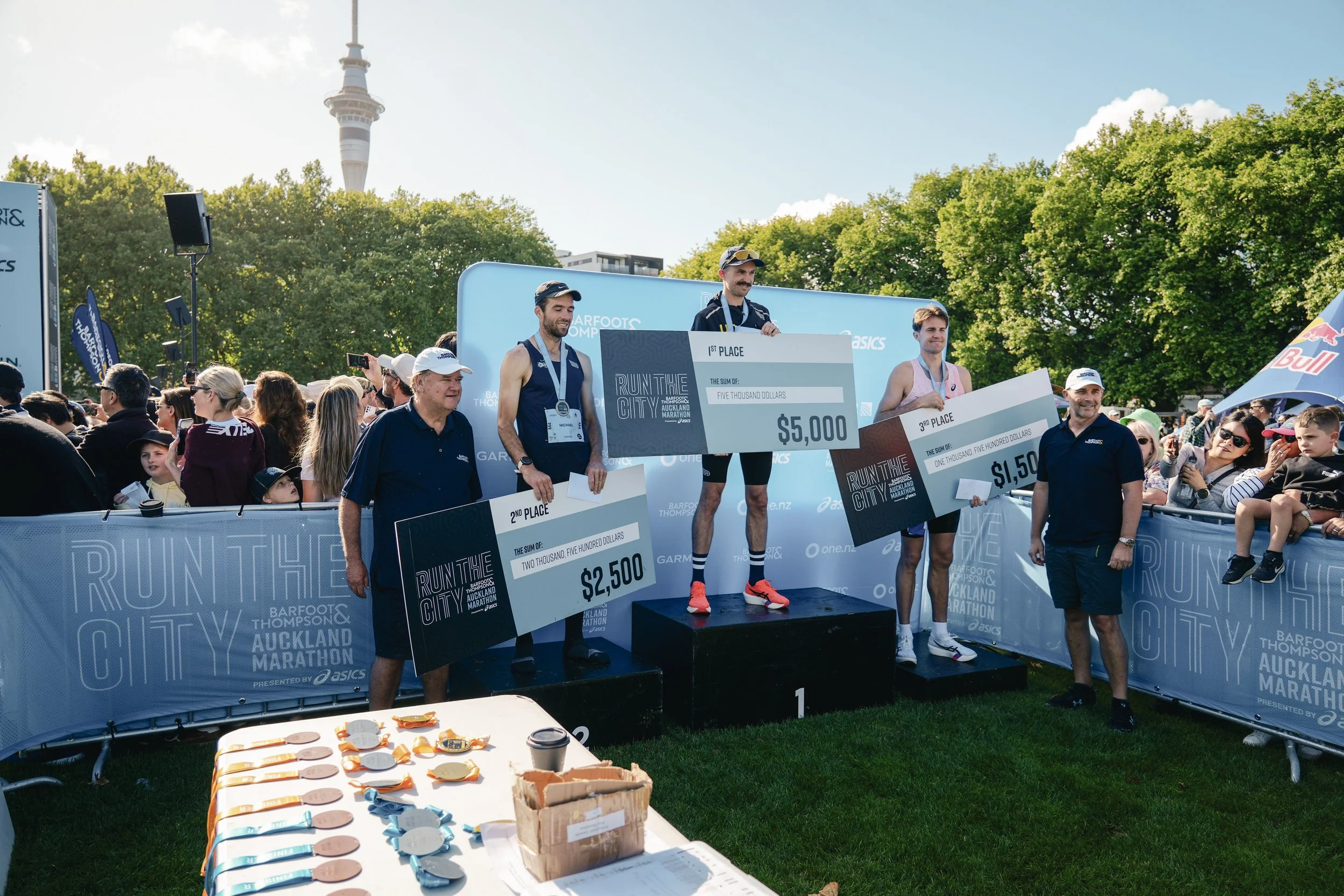 Marathon runners on a winners' podium with large checks in an outdoor park setting, crowd watching, and a TV tower in the background.