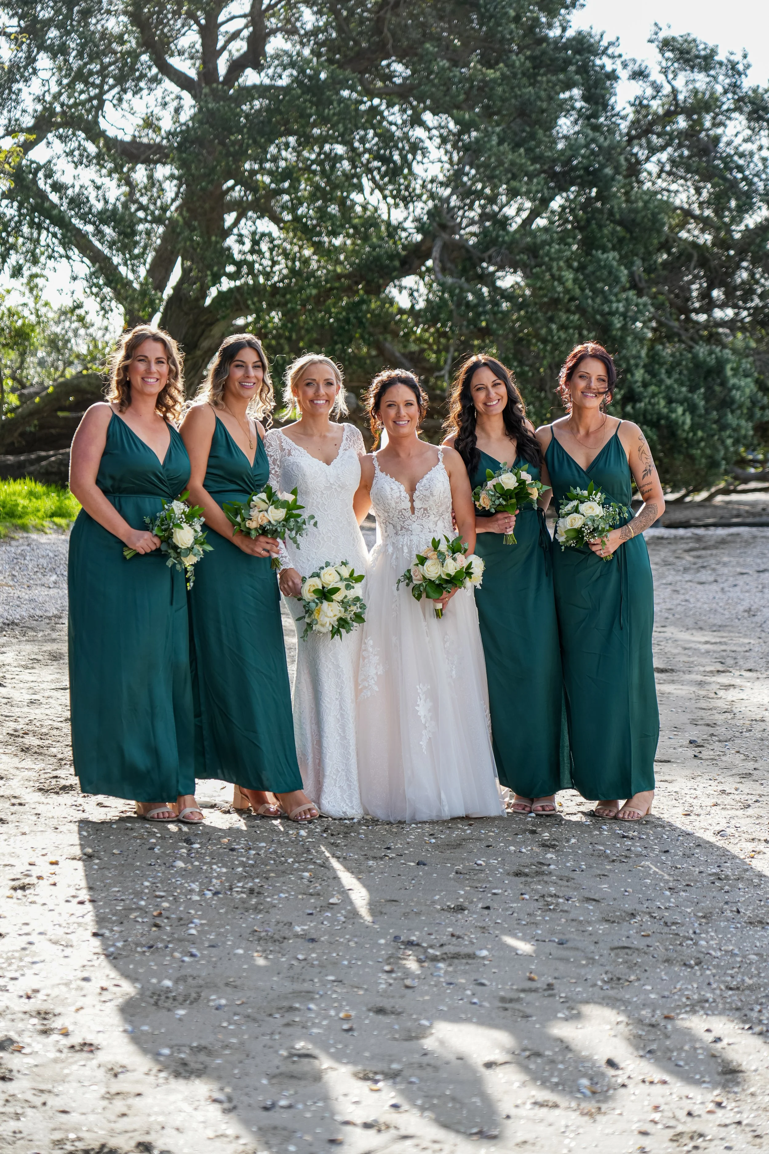 Bridal party of six women, two in white wedding dresses and four in green bridesmaid dresses, standing on a sandy beach with a large tree in the background, holding bouquets of white flowers.