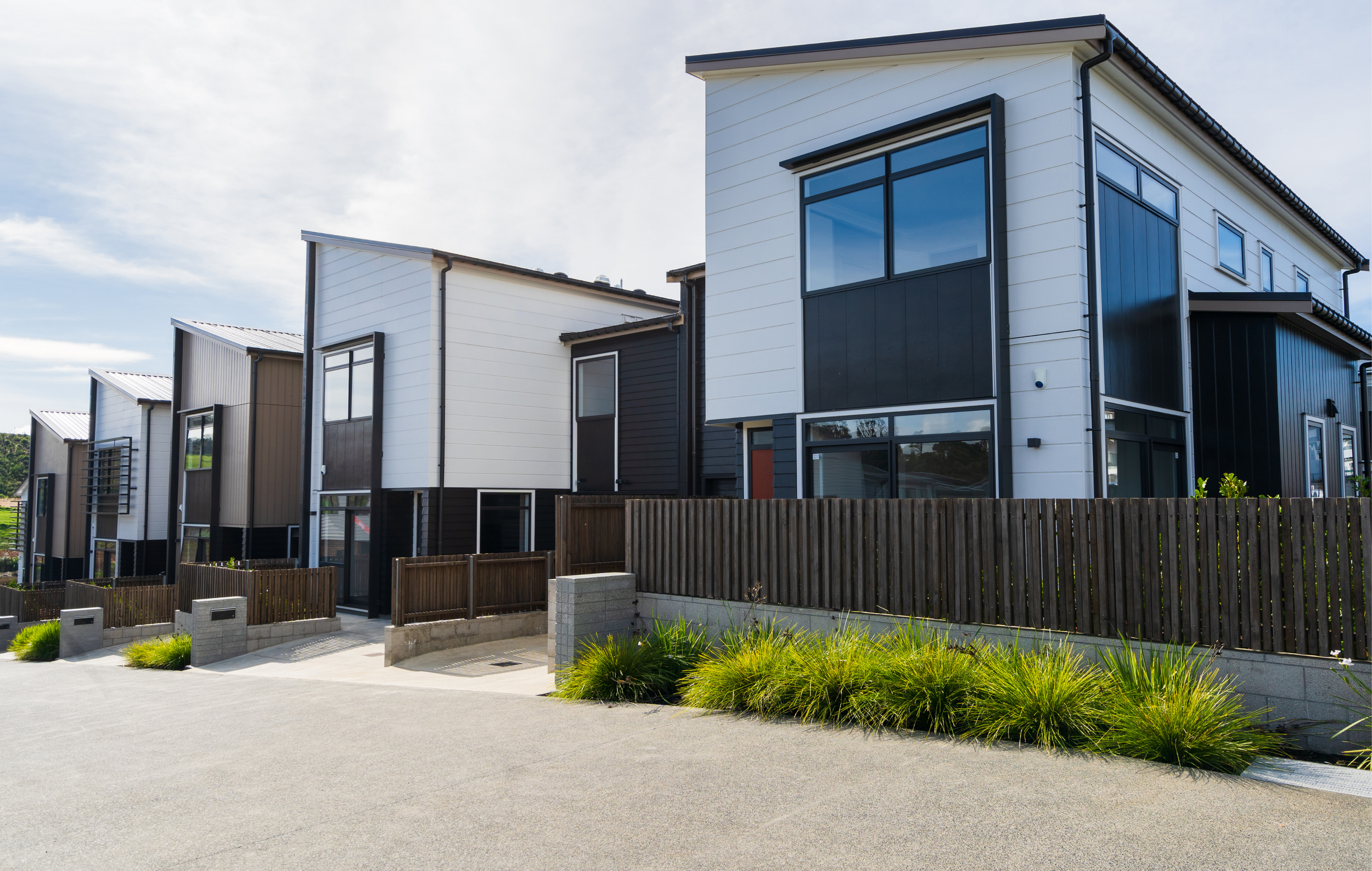 A row of modern two-story houses with white and dark exterior siding, large windows, a wooden fence, and landscaped greenery in the foreground.