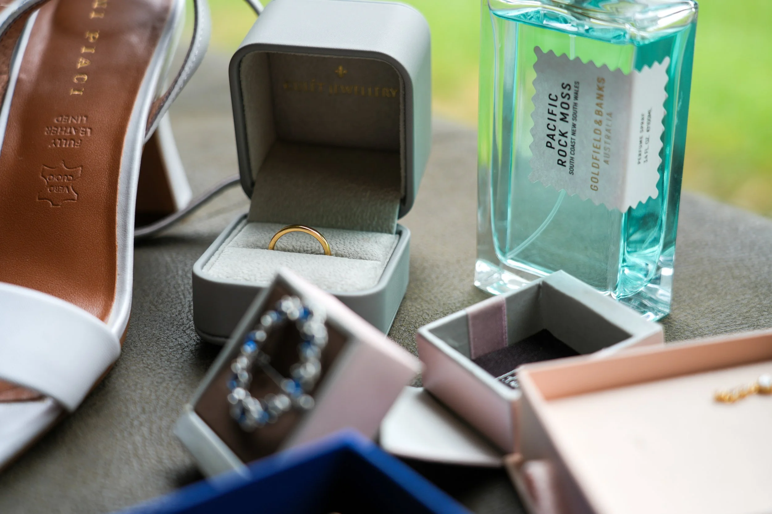 Various jewelry items in open and closed rings boxes, a pair of shoes with jewelry embossed on the sole, and a bottle of Pacific Rock Moss perfume on a beige fabric surface.