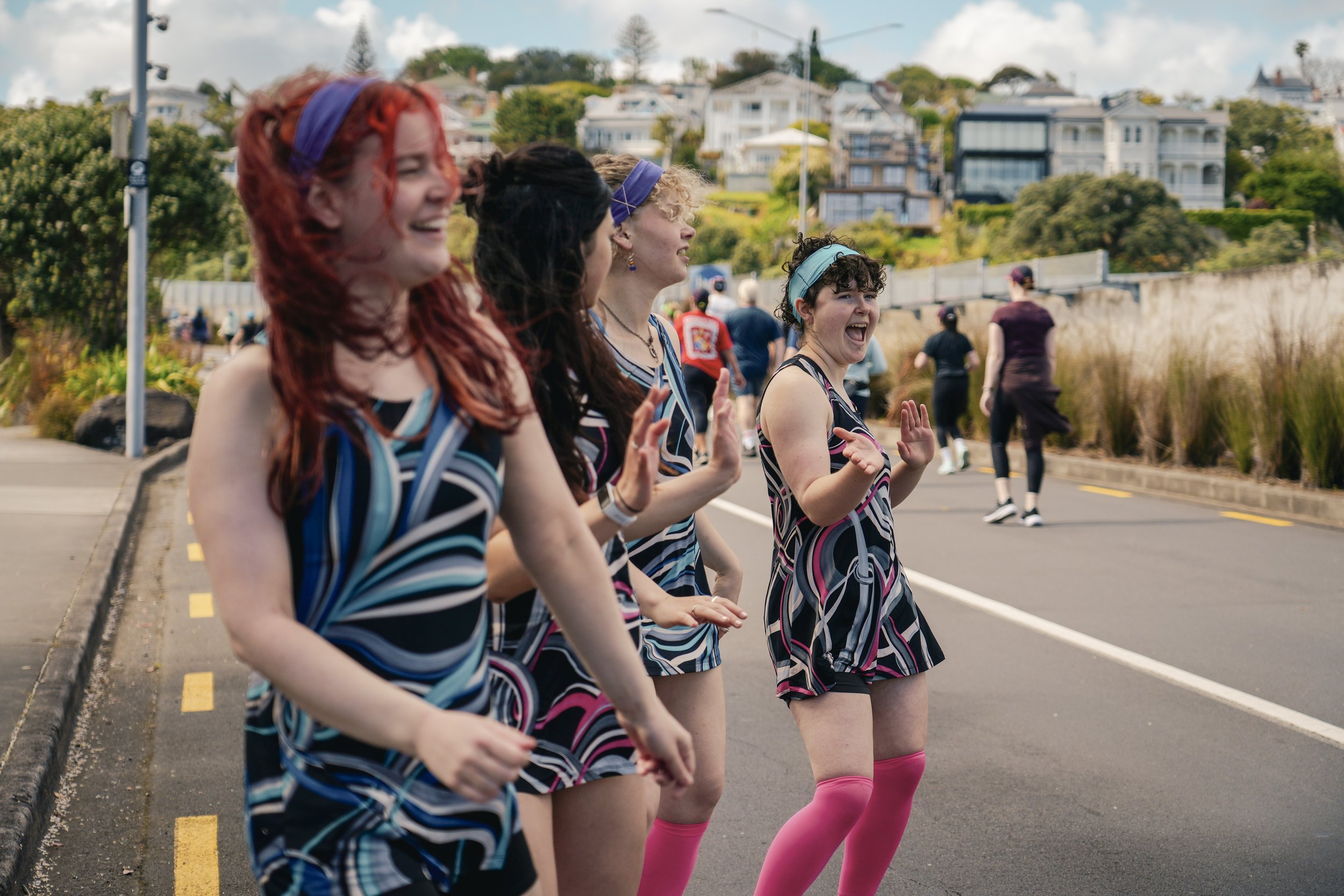 Four women wearing matching patterned dresses and pink knee-high socks standing on a street, smiling and waving at a parade or event. They are outdoors with houses and trees in the background.