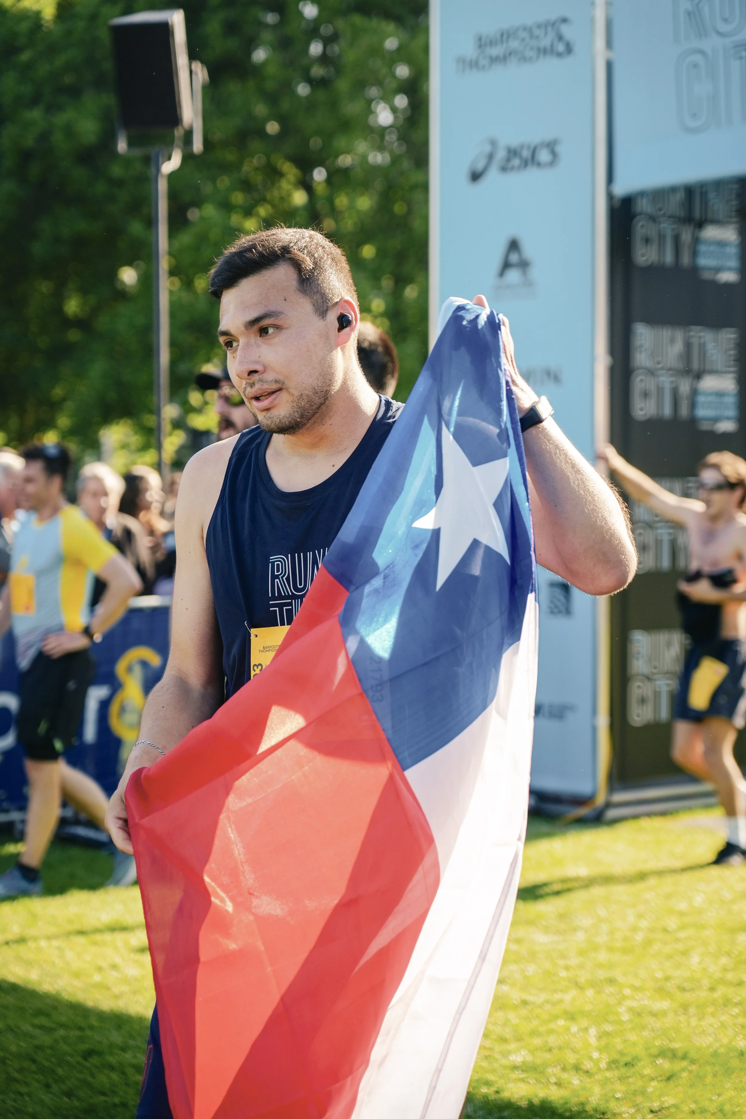 A man in running attire holding the Texas state flag after finishing a race at an outdoor event. There are other runners and onlookers in the background, and the scene is set in a park or green space.