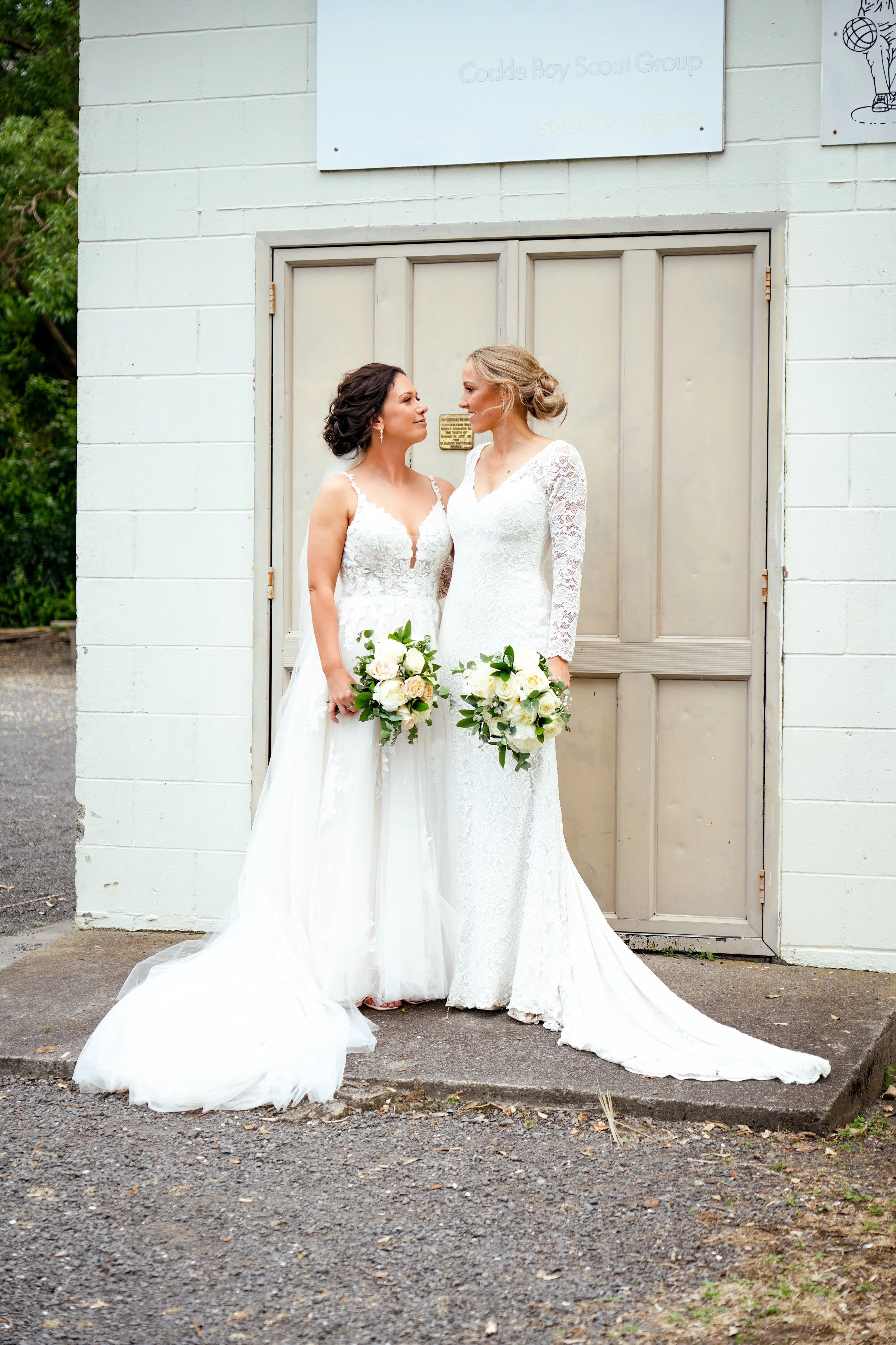 Two brides dressed in white wedding gowns looking at each other and holding bouquets of white roses and greenery, standing outside in front of a beige door and white brick wall.