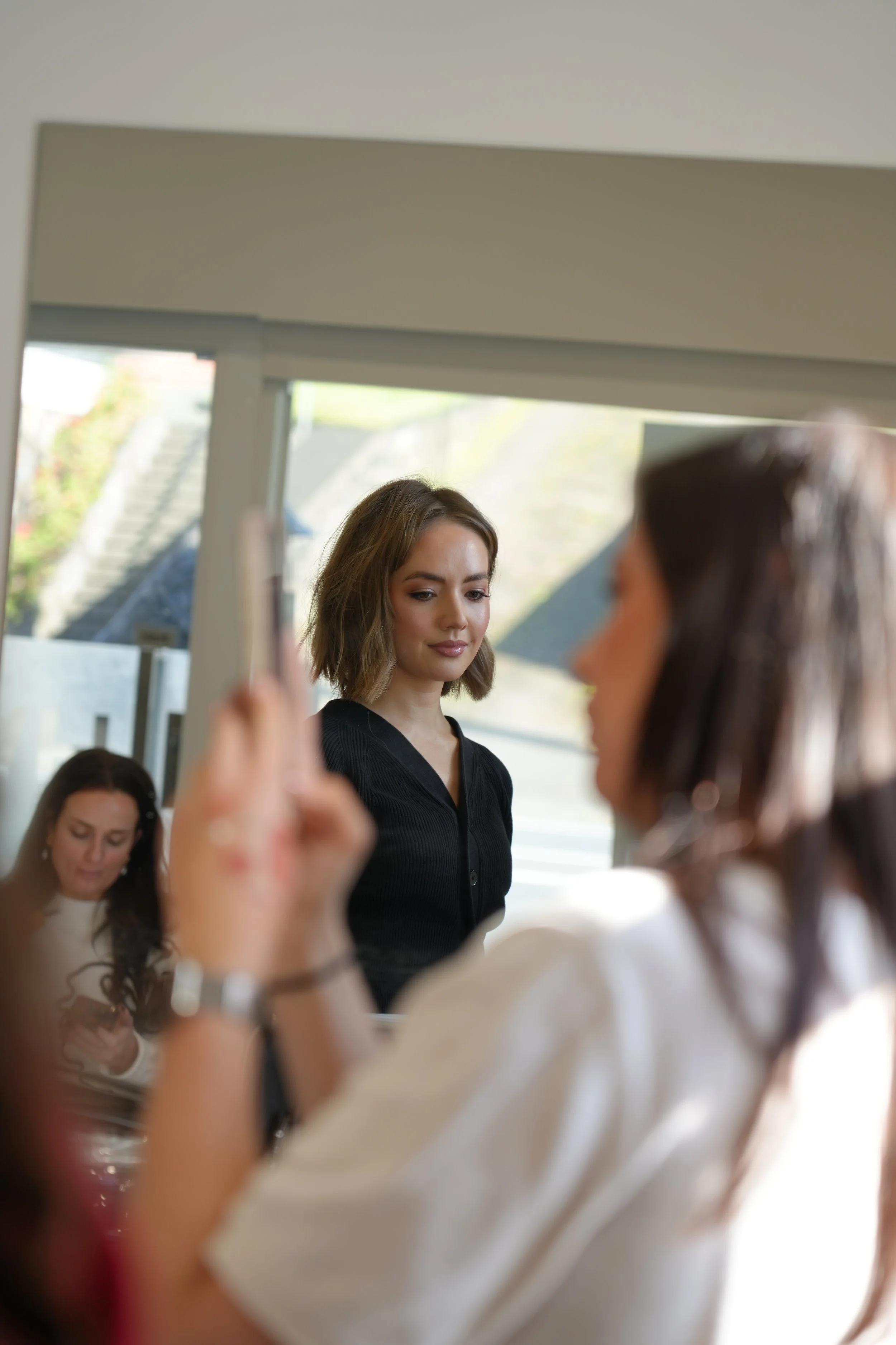 Women in a hair salon, one woman getting her hair styled and another woman taking a photo with her phone.