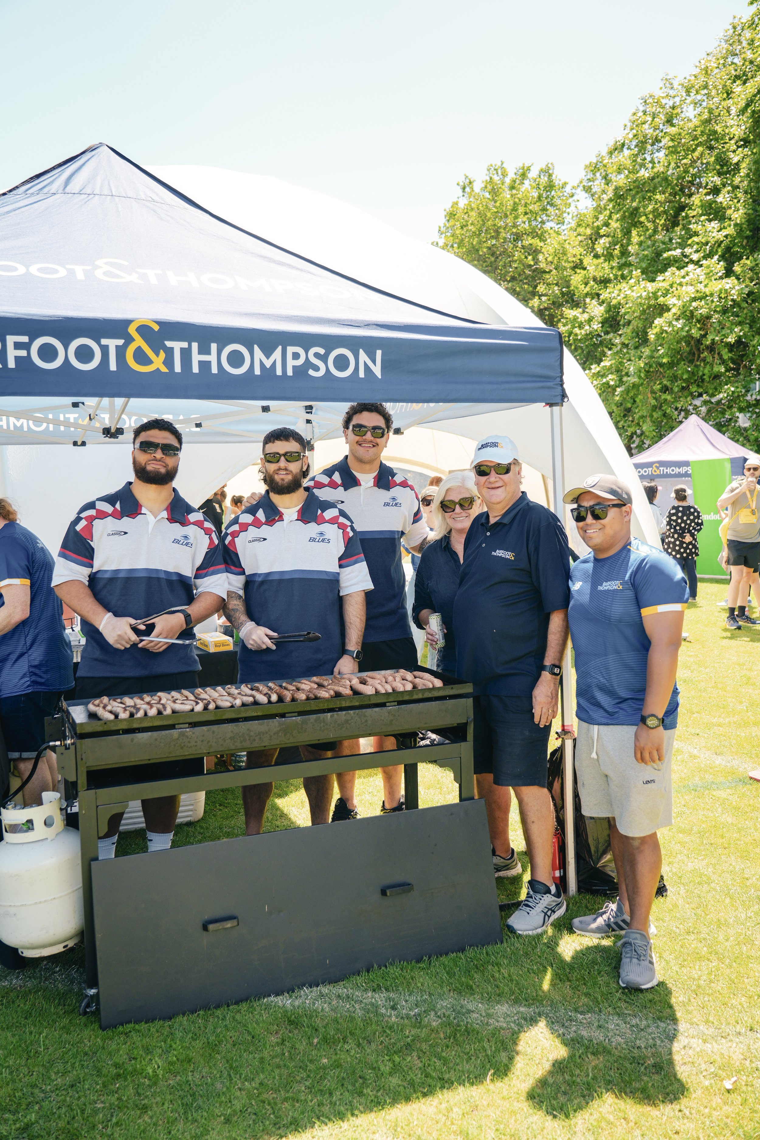 Group of people smiling at a grilling event outdoors, with a blue and white canopy displaying 'FOOT & THOMPSON'