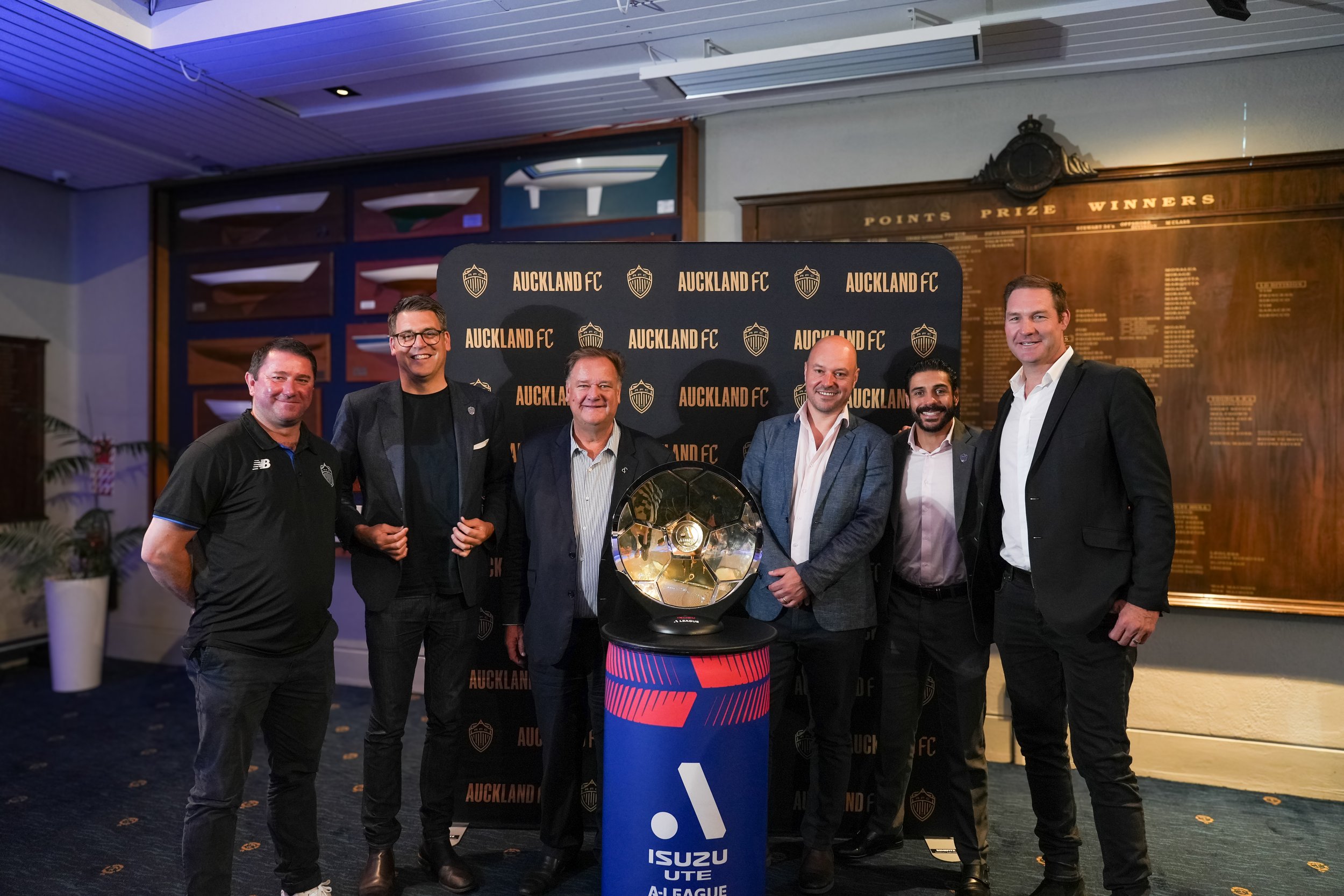 Group of seven men standing around a trophy at an Auckland FC event, with a backdrop displaying the club's name, and a wall with memorabilia and a points and winners board in the background.