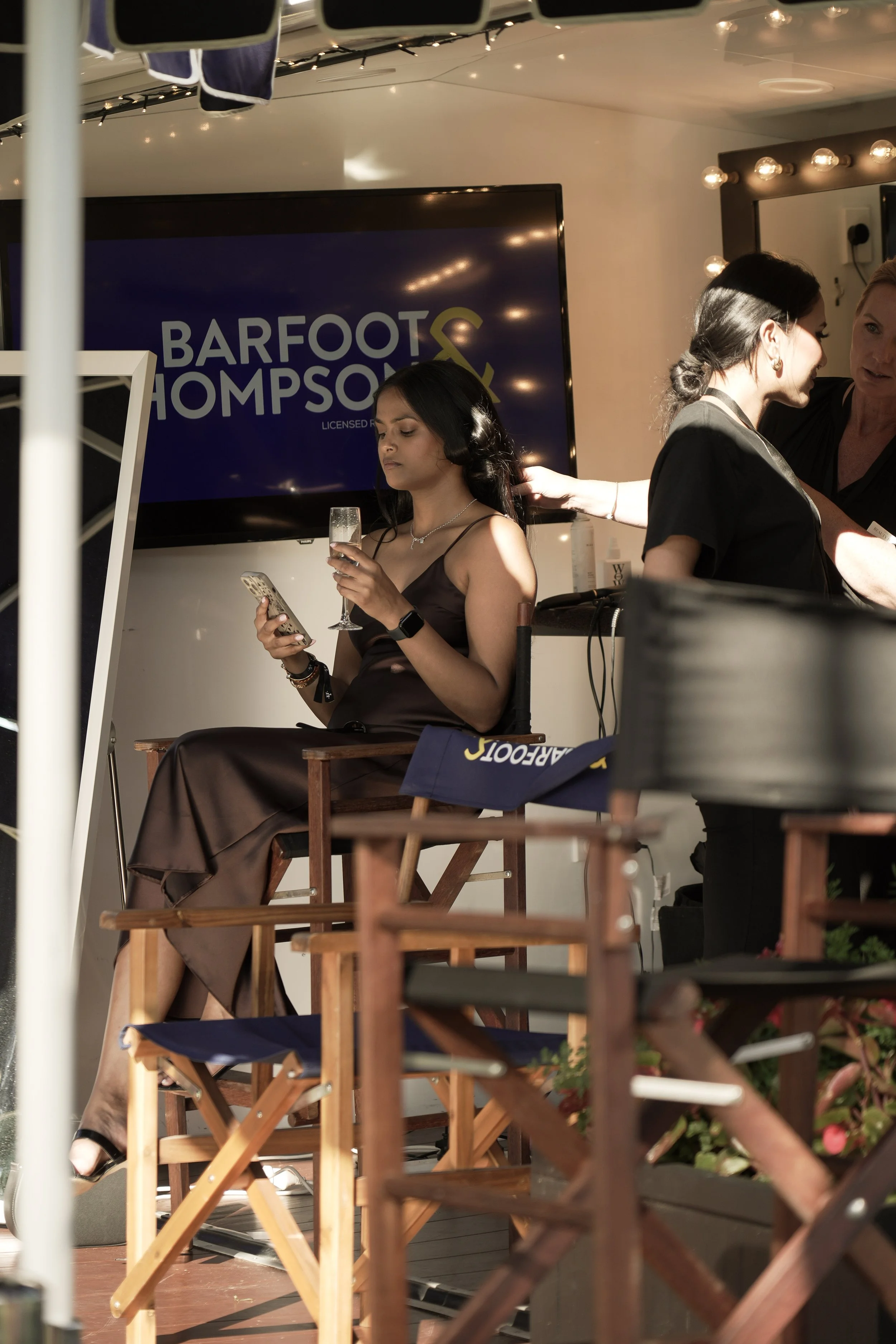 A woman with long dark hair wearing a black dress sitting on a wooden chair, holding a glass and looking at her phone. Behind her, two women are styling hair. In the background, a television screen displays the text "BARFOOT & THOMPSON".