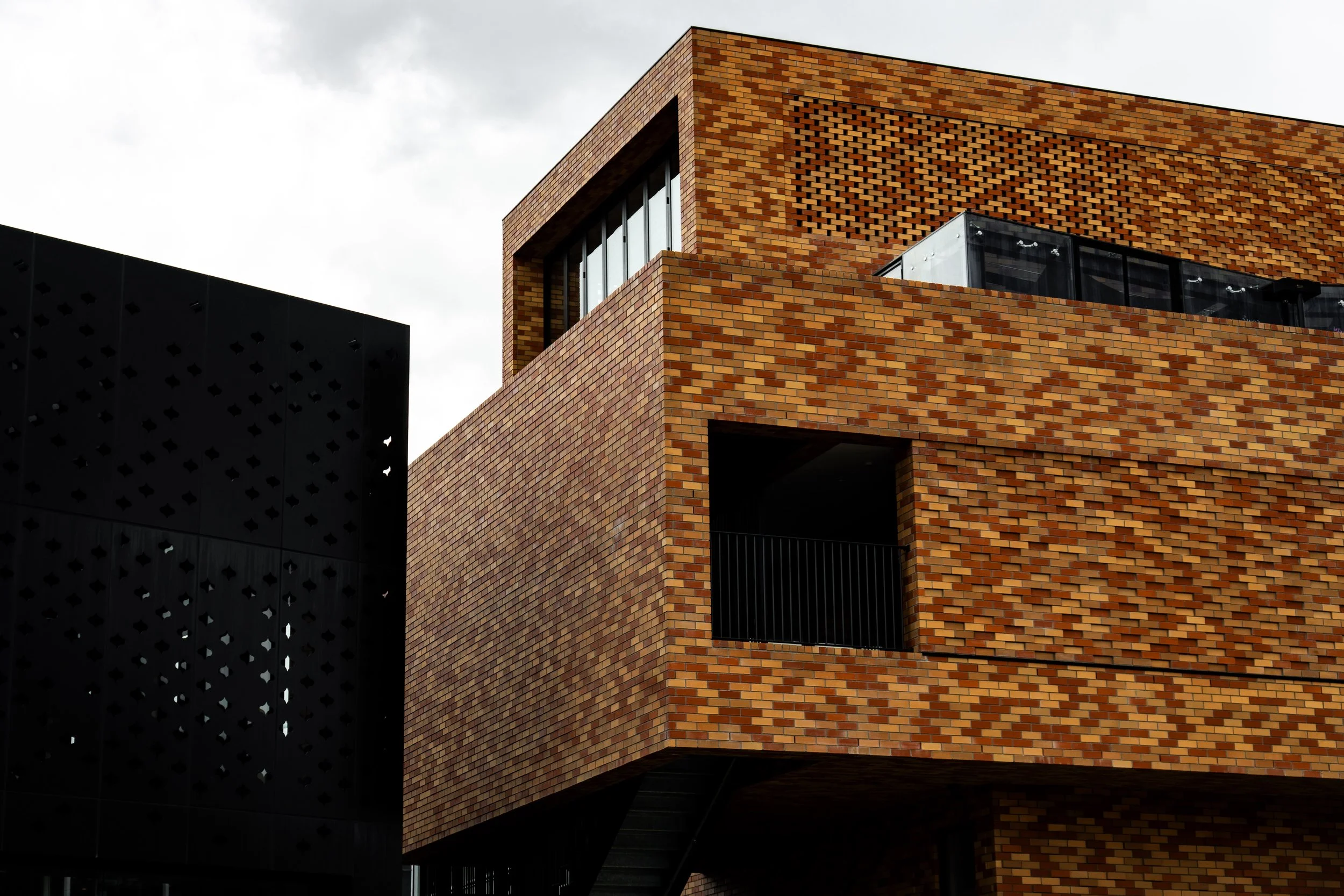 Close-up of a modern building with a brick facade, featuring large windows and a balcony, with part of a black metal structure in the foreground.