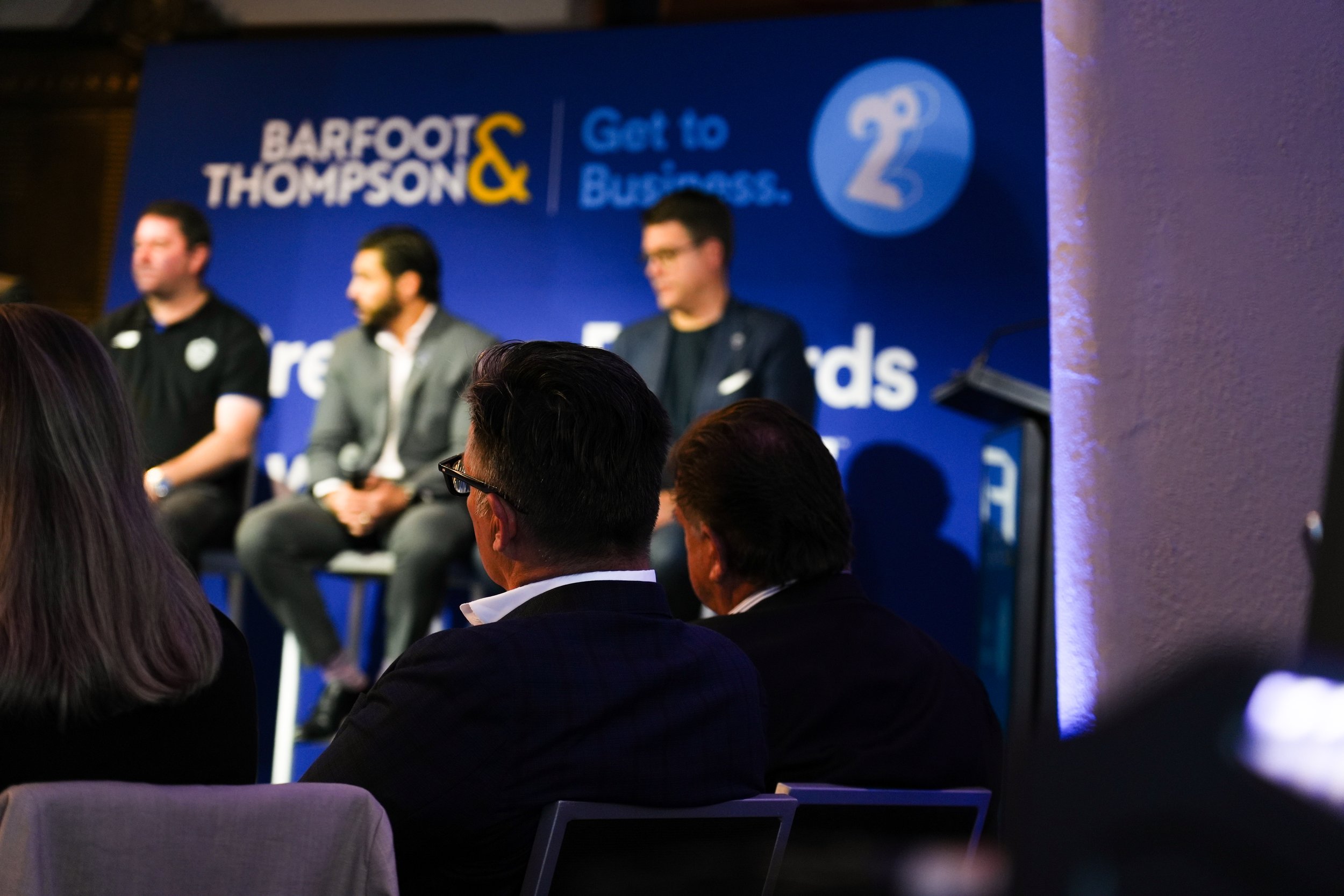 People seated at a panel discussion at a conference with a blue backdrop displaying the logos and text for 'Barefoot & Thompson' and 'Get to Business'.