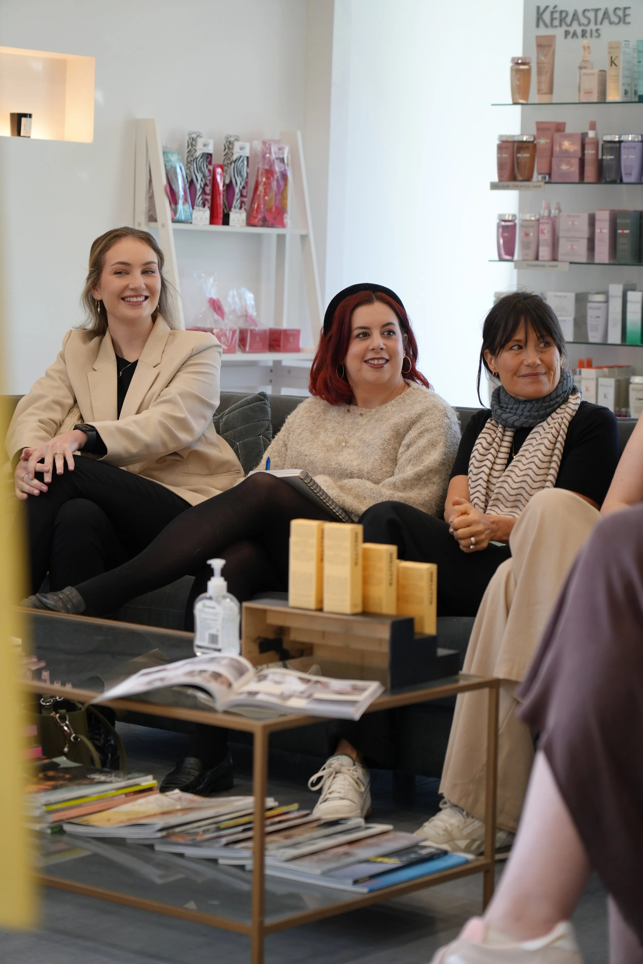 Group of women sitting on couch in a store or salon, smiling and engaged in conversation, with shelves of hair or beauty products in the background.