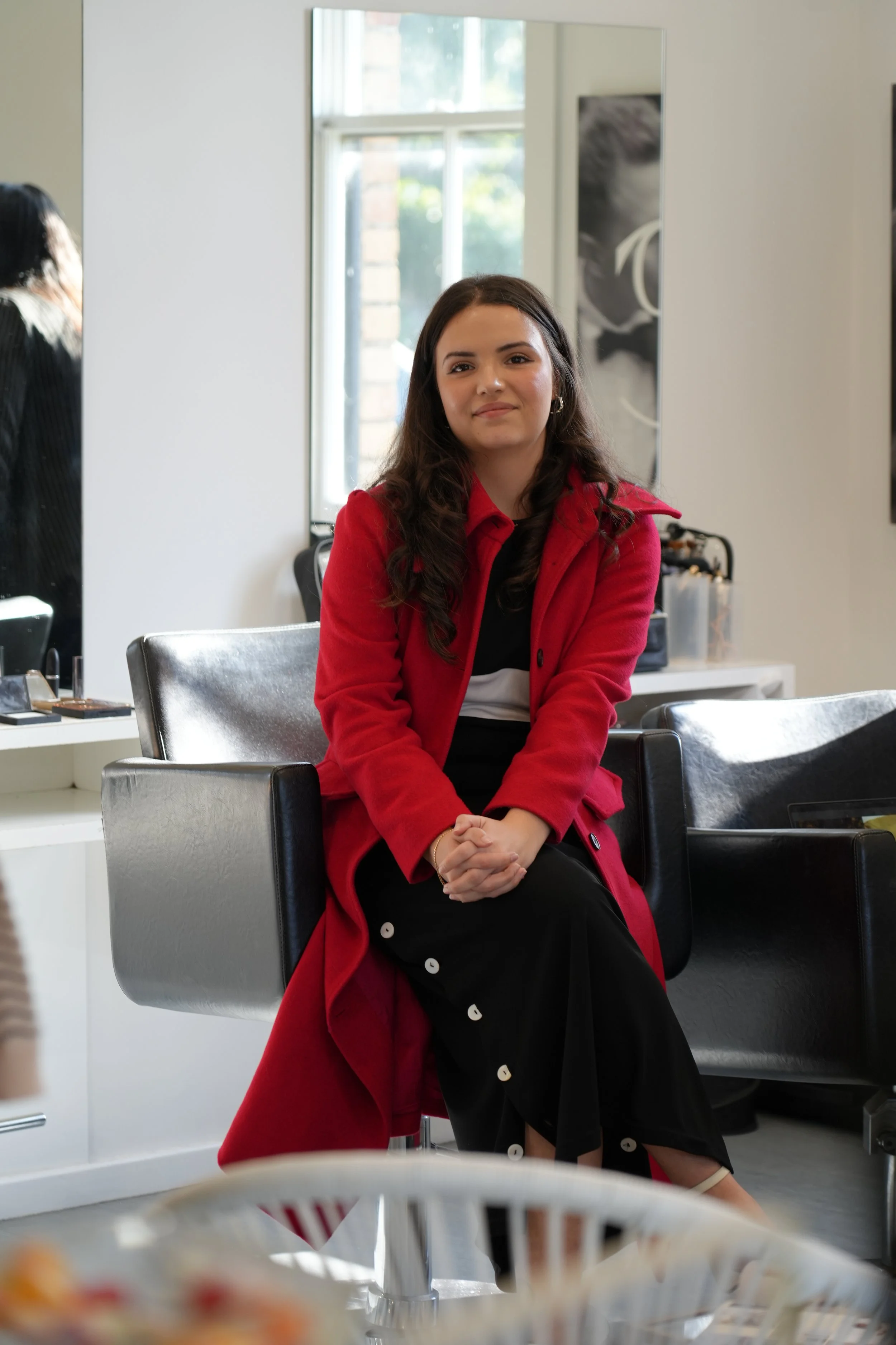 A young woman with long dark curly hair, wearing a red coat, black and white outfit, sitting on a black salon chair inside a hair salon with a mirror behind her and styling tools on a counter.