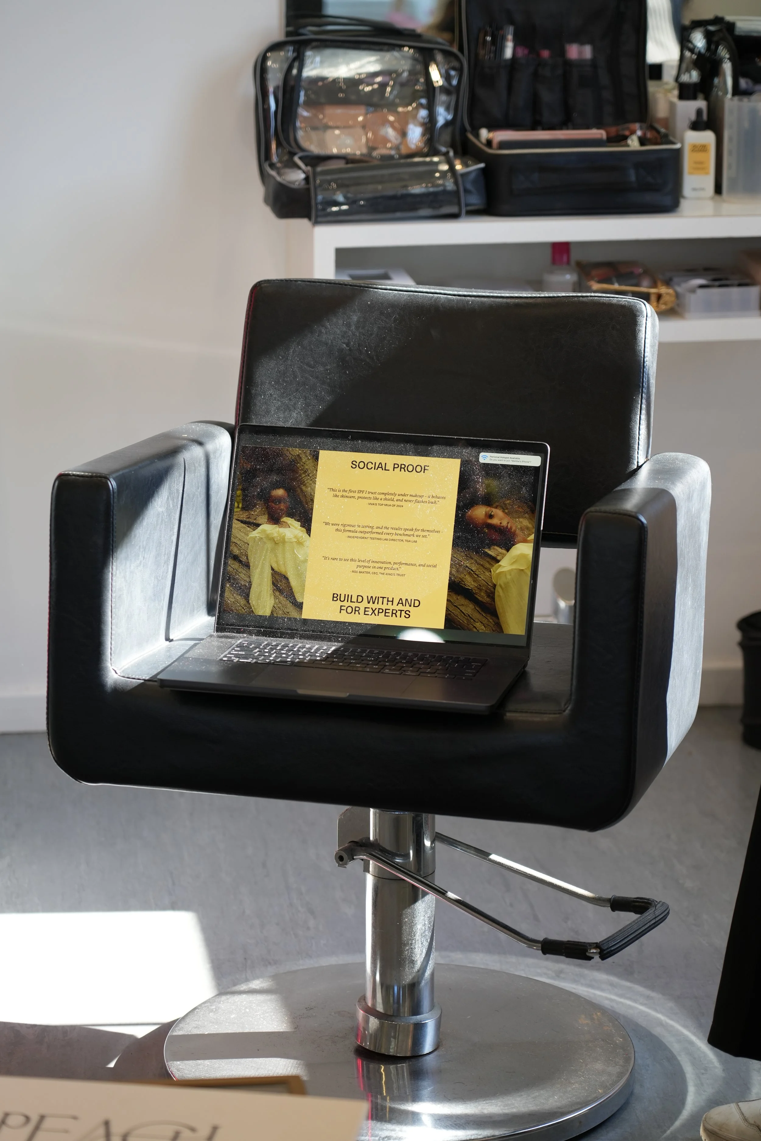 A black salon chair with a laptop showing a presentation titled 'Social Proof' on the seat. In the background, there is a white shelf and various makeup and beauty supplies.