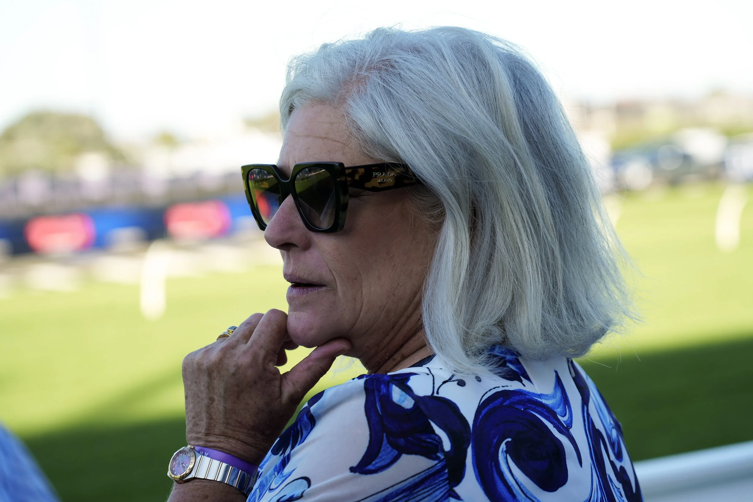 A woman with gray hair wearing black Prada sunglasses with tortoiseshell arms, a gold watch, and a blue and white patterned top, sitting outdoors on a grassy field with blurred background of trees and parked cars.