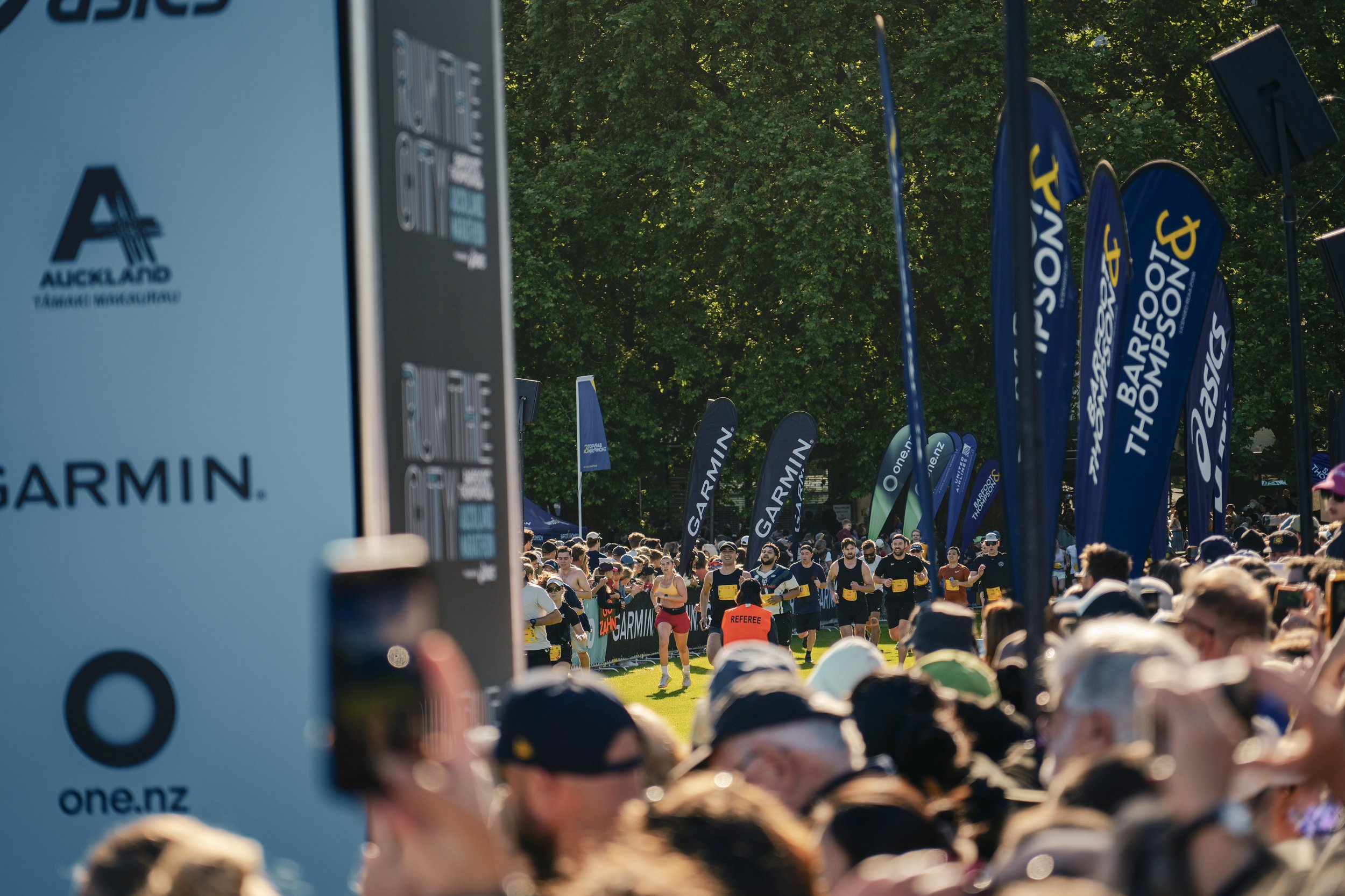 Crowd of runners at start line of a race organized by Auckland and Garmin, with flags and banners in the background, outdoors on a sunny day.