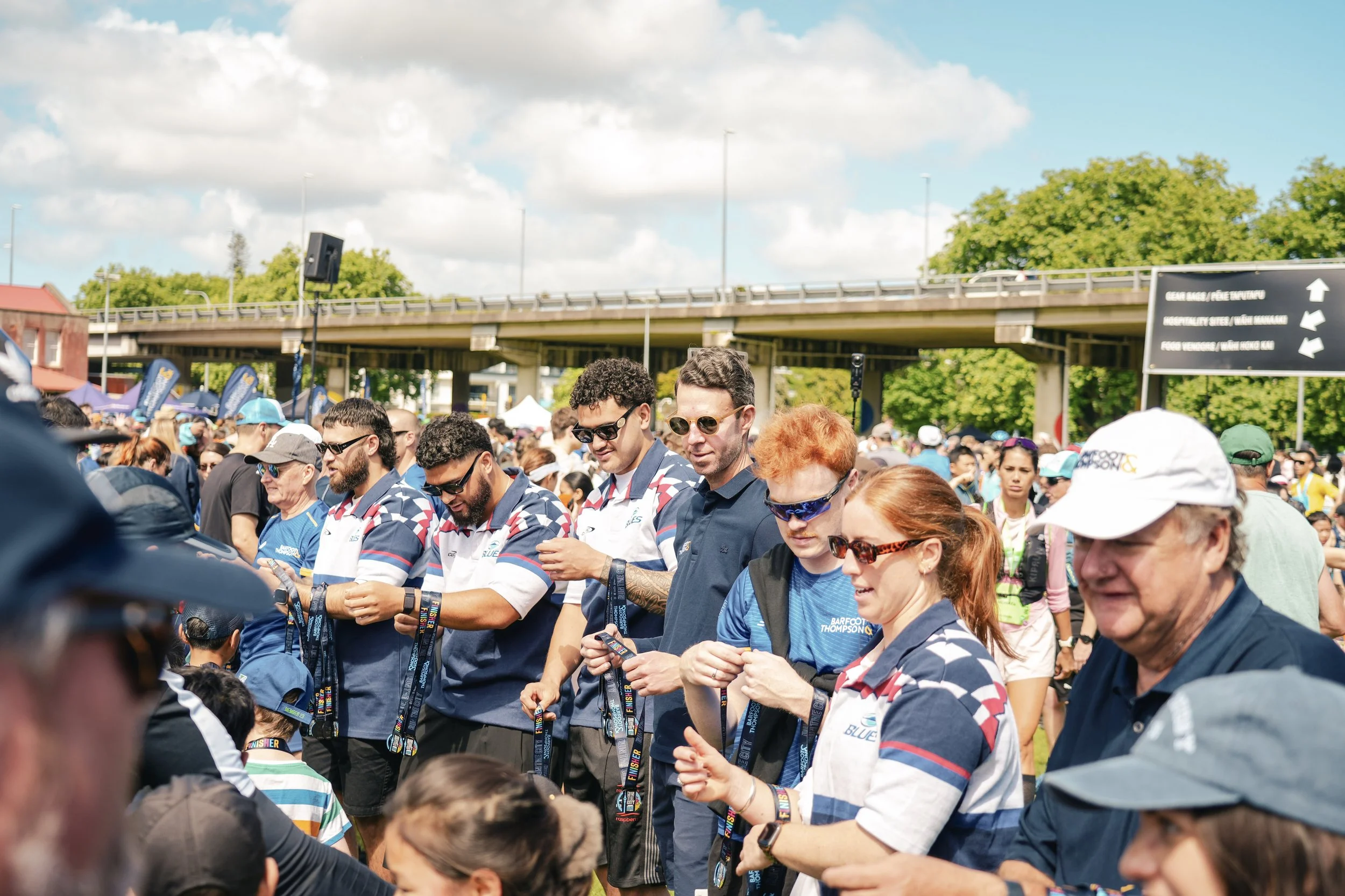 Crowd of people gathered outdoors for an event, many are wearing sunglasses and holding medals, with a bridge and trees in the background on a sunny day.