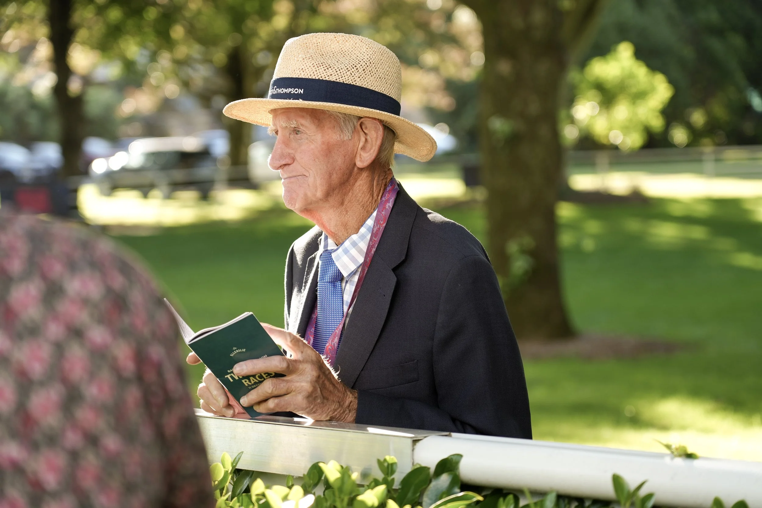 An elderly man wearing a straw hat and a dark blazer is holding a small booklet while standing outdoors near a white fence, with trees and parked cars in the background.