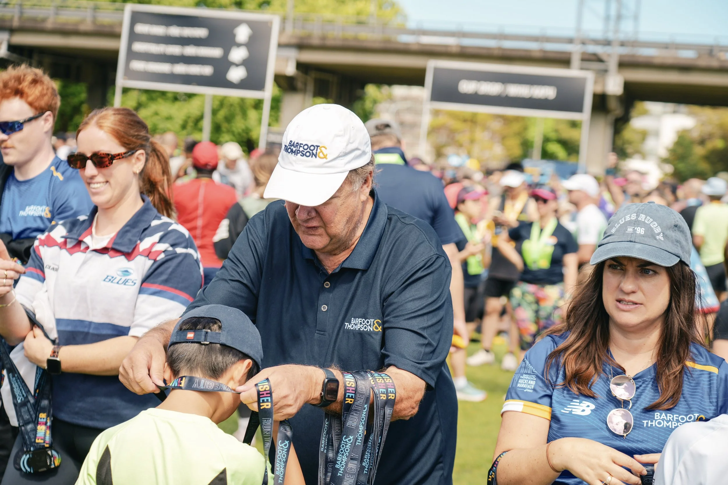 Group of people gathered at a race event, with a man awarding a young girl with medals on a sunny day, some wearing event shirts and hats, amid a crowd with banners in the background.