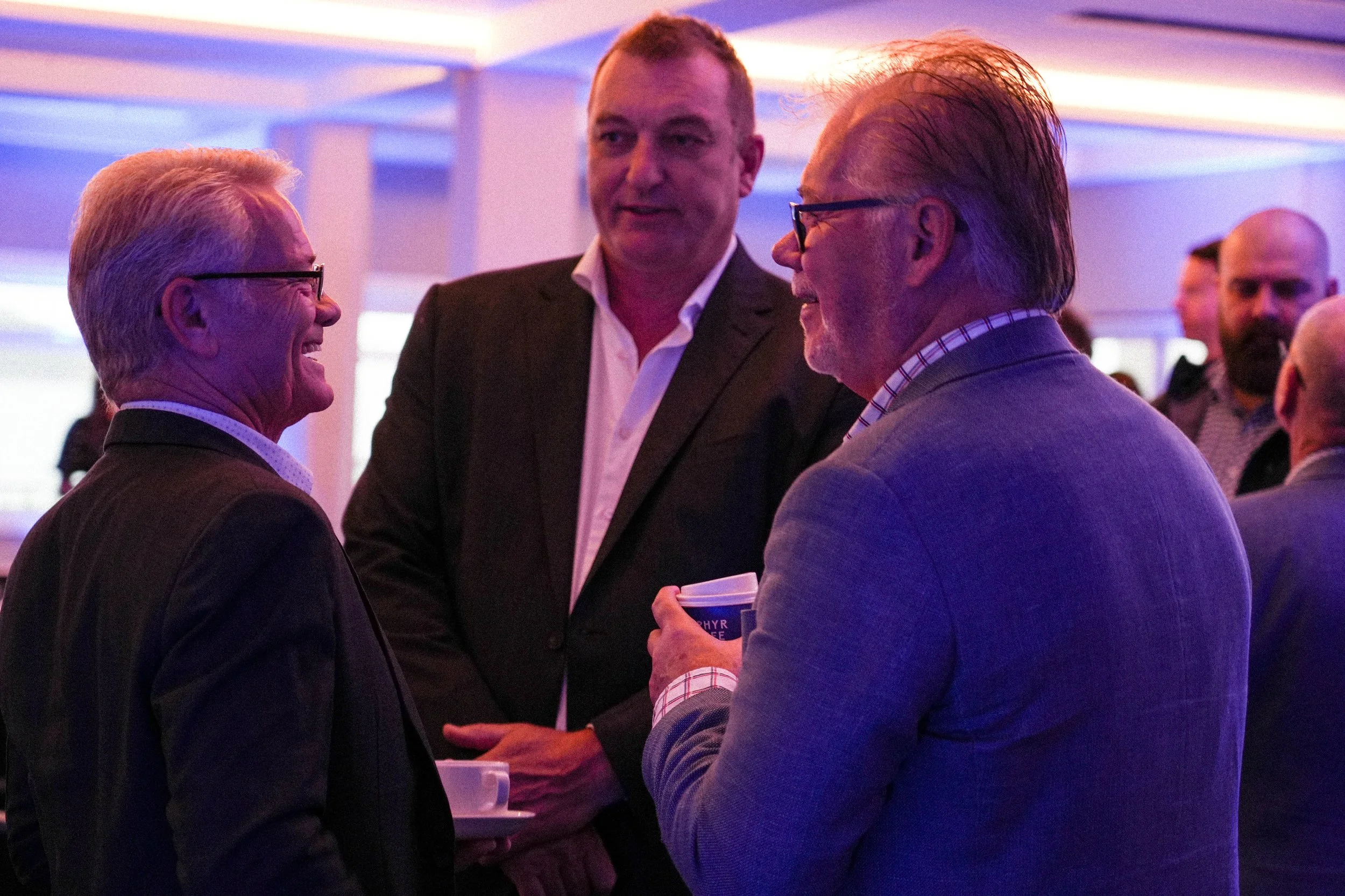 Three men in conversation at a professional event with purple and pink lighting.