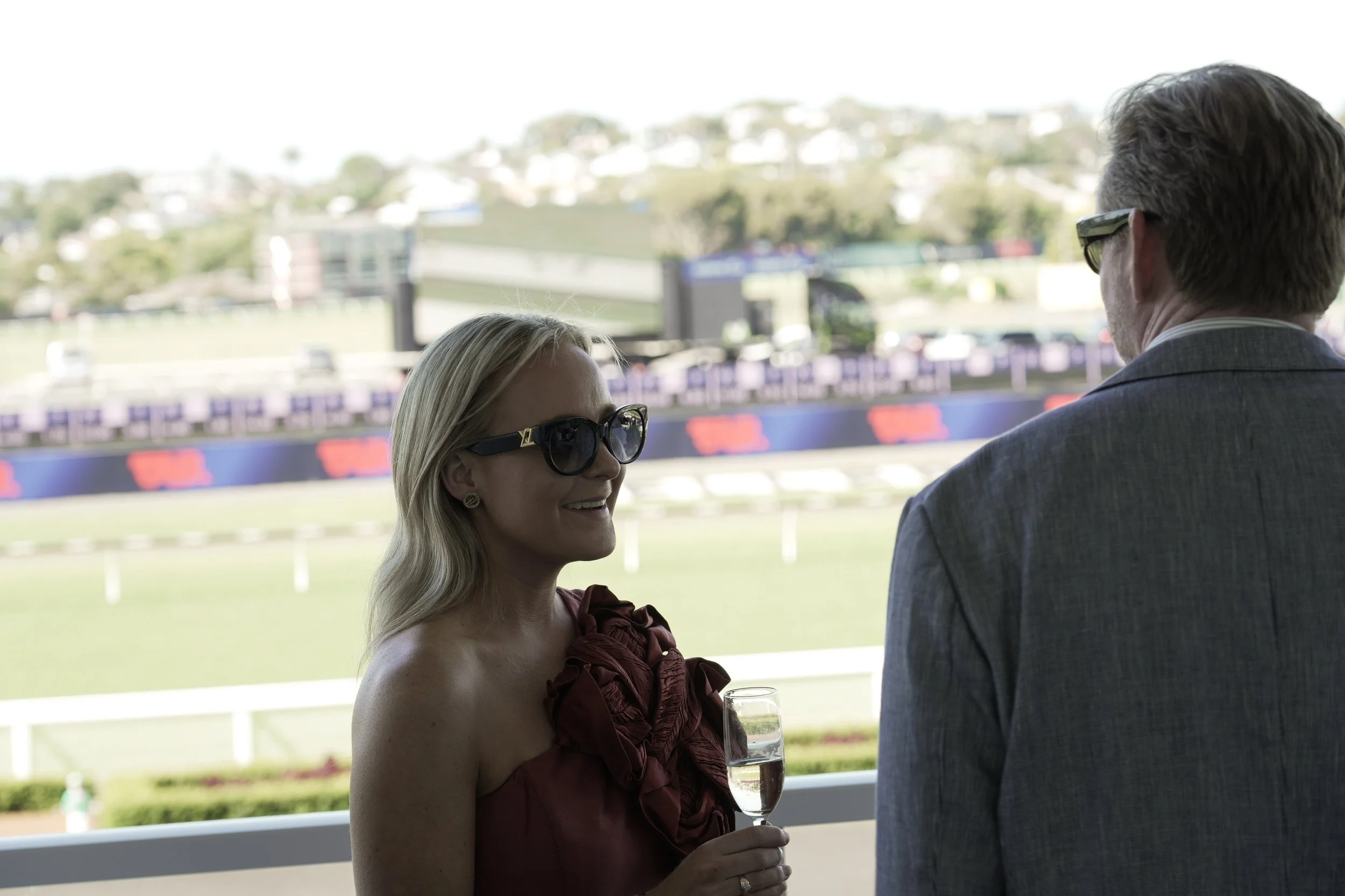 A woman wearing sunglasses and a strapless dress holding a glass of champagne, conversing with a man in a suit at a horse racing event, with a racetrack and hills in the background.