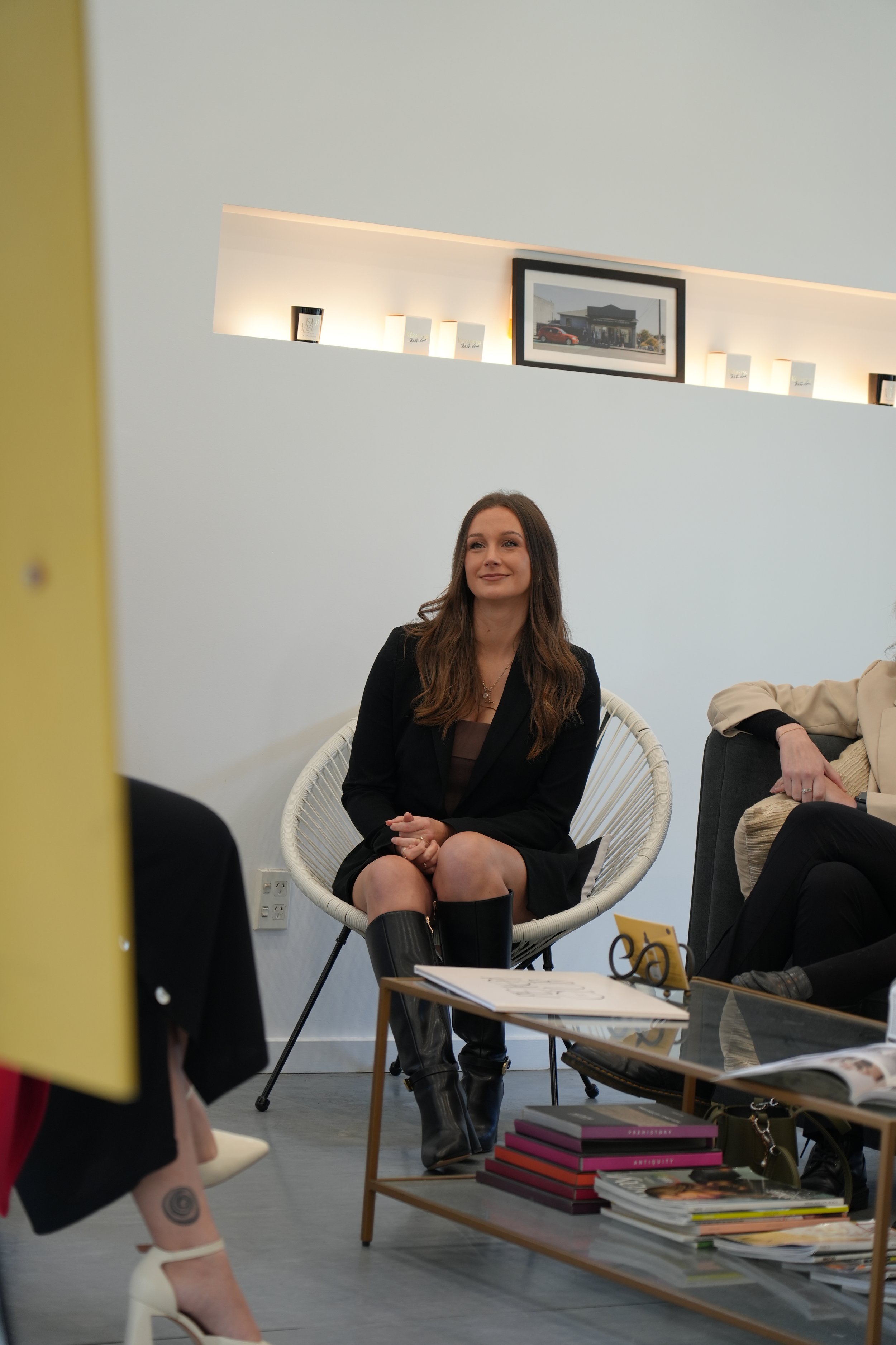 A woman sitting on a white, round chair with black boots and a black blazer, smiling during a discussion in a modern, brightly lit room with a gallery wall.