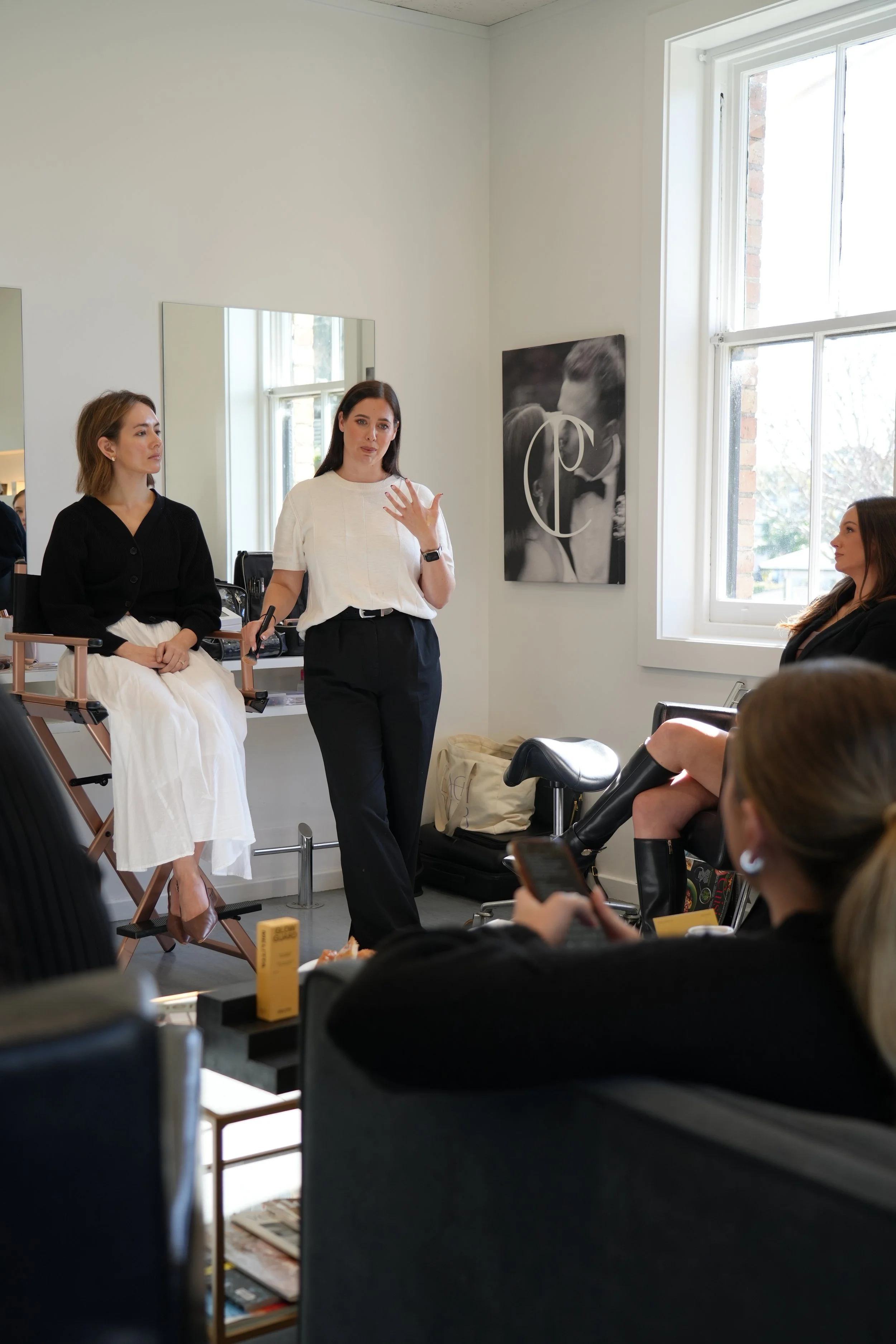 A woman in white with black pants is speaking to an audience in a room with white walls and large windows. Two women are seated, one with a black top and the other with a black dress and boots, listening attentively.